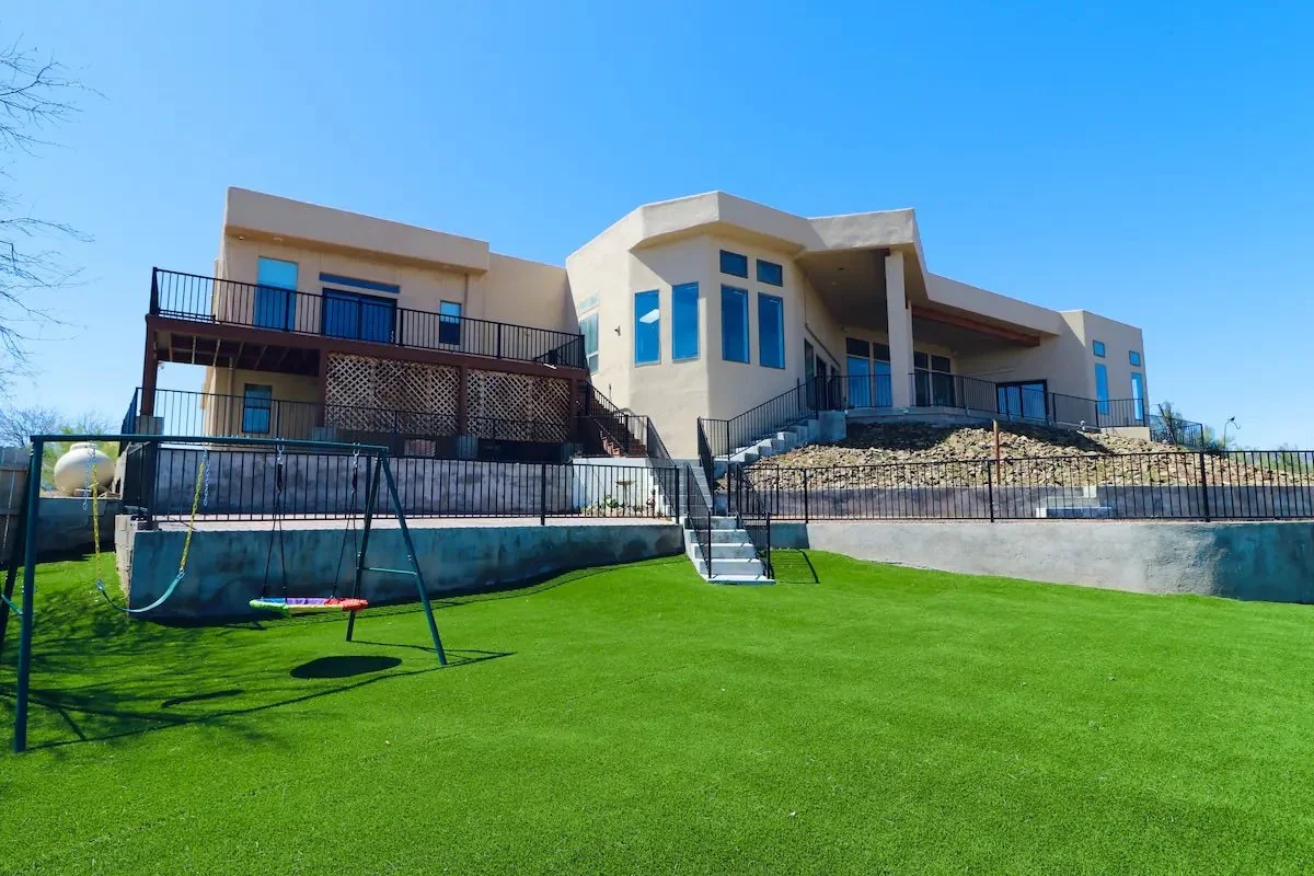 Modern house with sloped roof, large windows, and multiple terraces, situated on a grassy backyard with a swing set and fence.