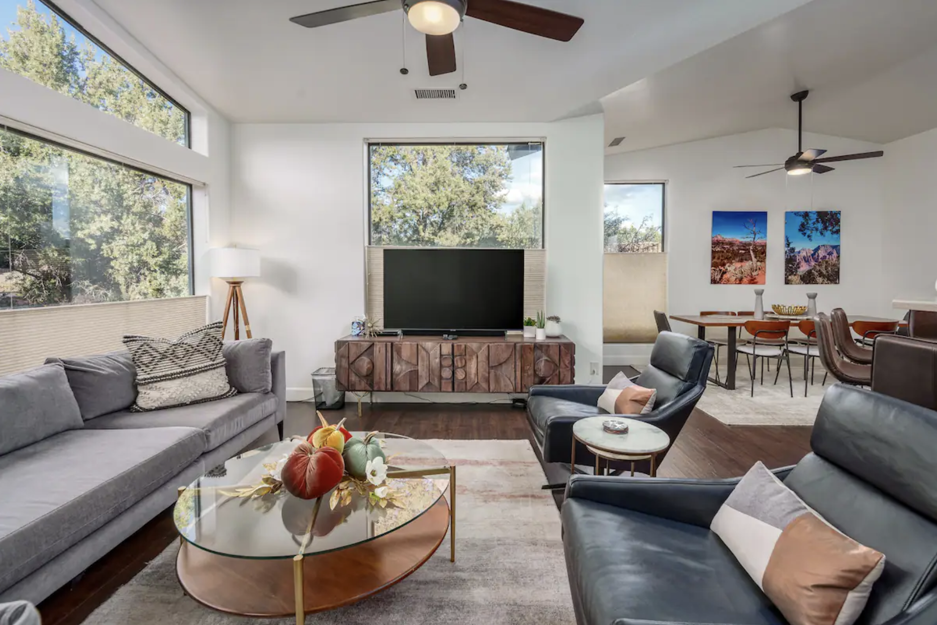 Modern living room with gray sofa, black leather chairs, a round glass coffee table with decorative pumpkins, a wooden media console with a TV, large windows with outdoor trees, and a dining area with abstract landscape art on the wall.