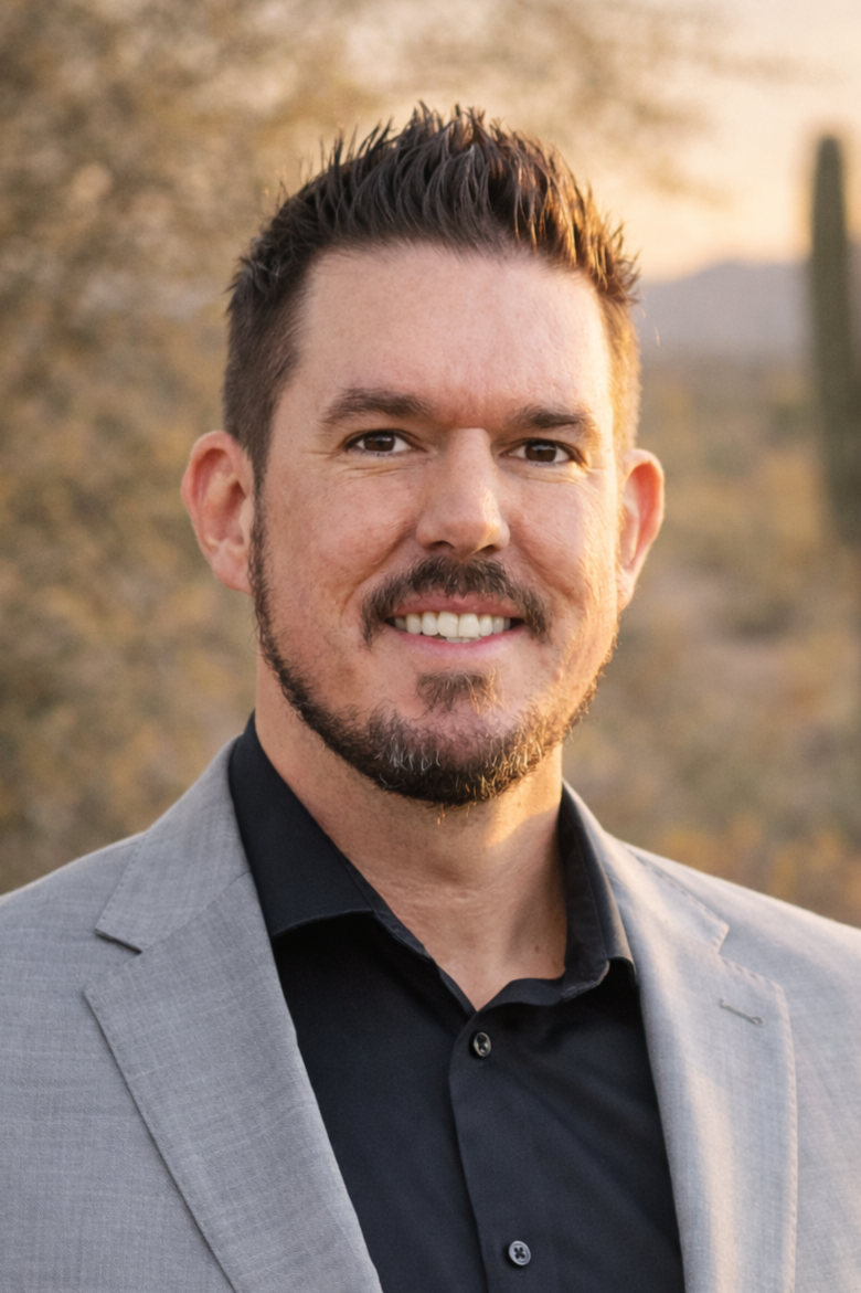 A professional headshot of a man with dark hair, a beard, and mustache, wearing a blue shirt and a black blazer, smiling against a dark background.