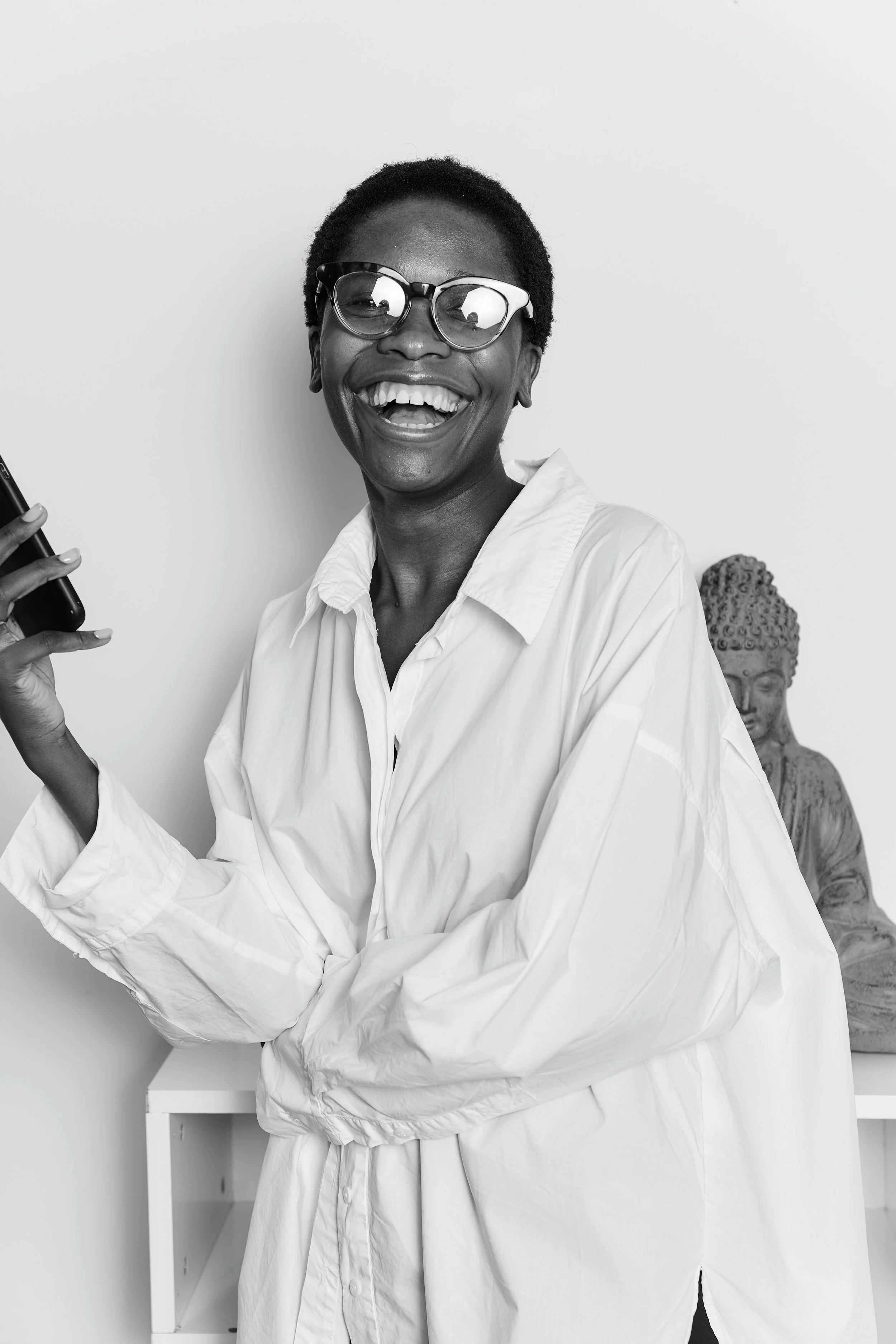 A smiling Black woman wearing glasses and a white button-up shirt, holding a phone, standing near a table with a Buddha statue in the background, indoors.