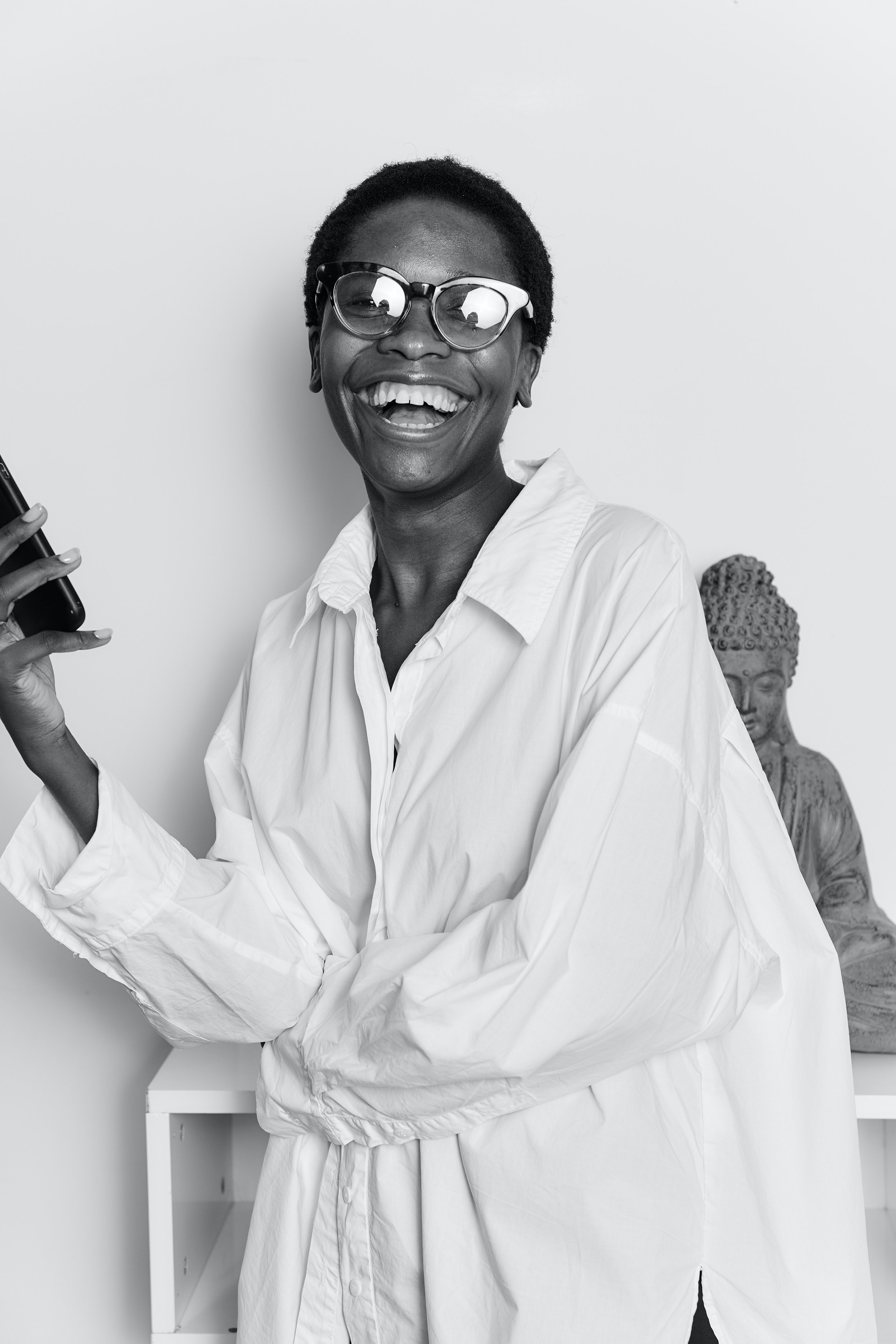 A woman with short hair and glasses, smiling and holding a phone in her right hand, standing against a white wall with a Buddha statue in the background.