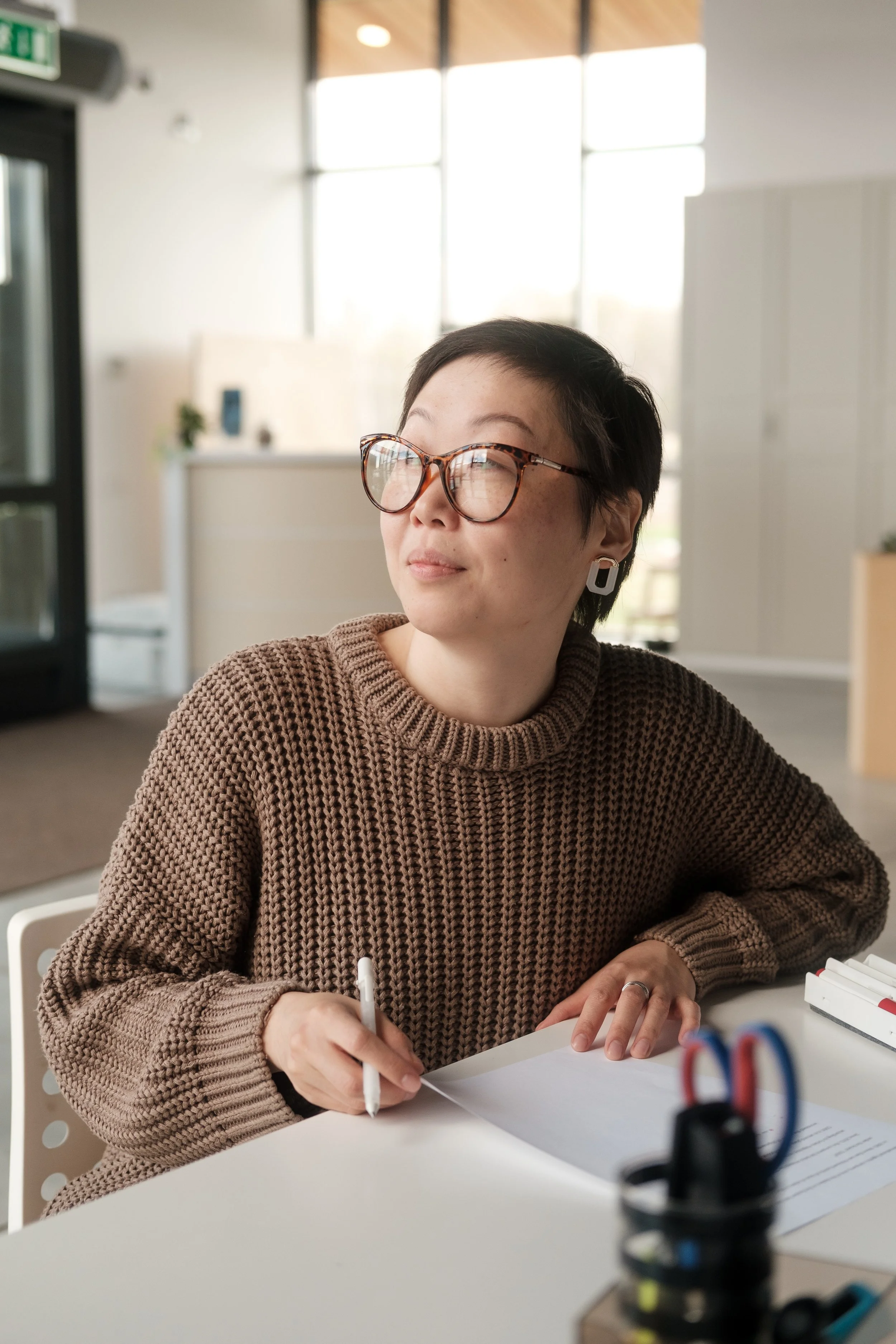 A woman with short dark hair and glasses sits at a desk, writing on a piece of paper, wearing a brown knit sweater and earrings in a well-lit office with large windows.