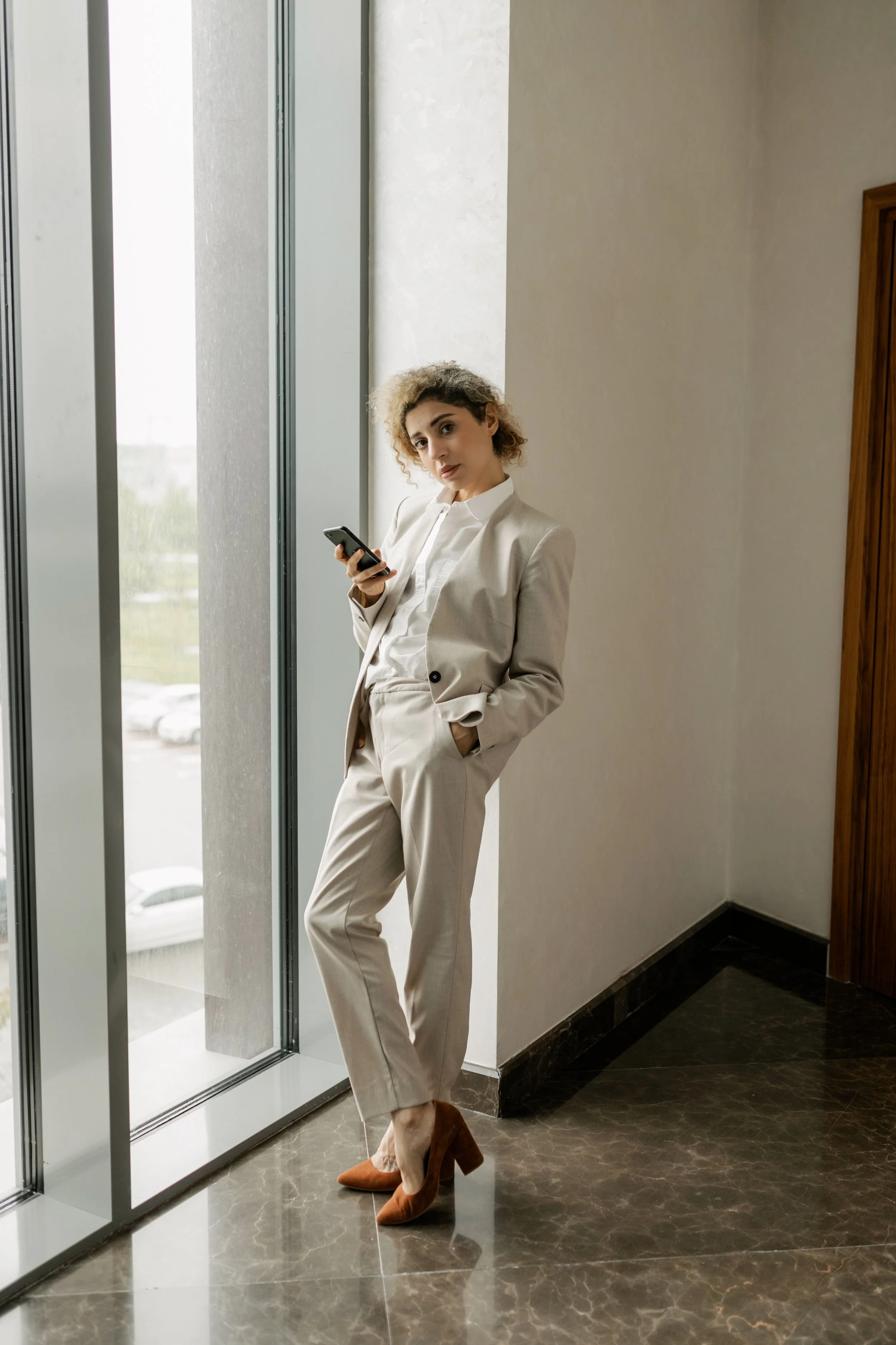 A woman in a beige pantsuit and brown high heels stands near a large window, looking at her phone, inside a modern office building.