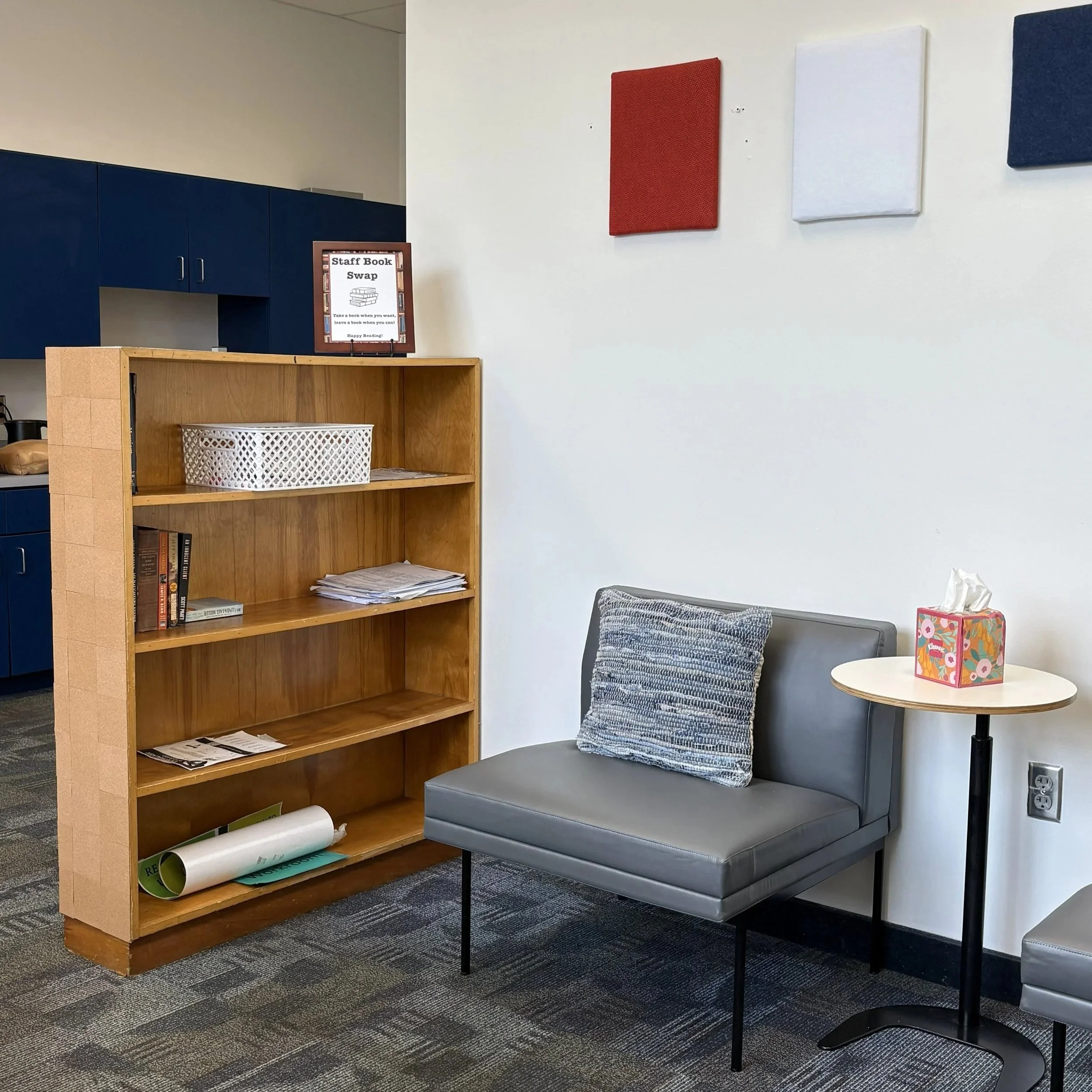 A kitchenette area with a white refrigerator, black microwave ovens, a white sink, coffee maker, and blue cabinets against a white brick wall.