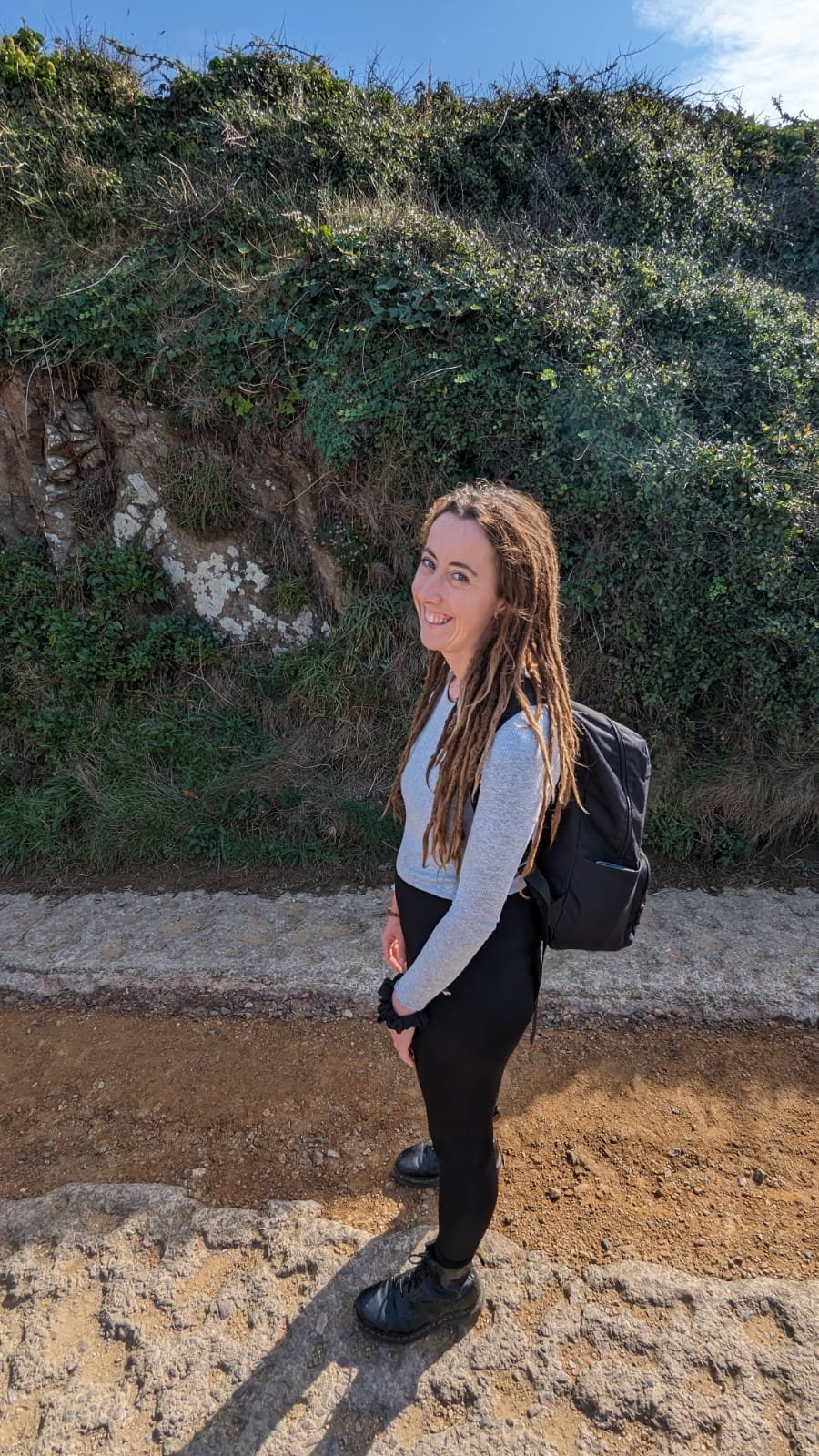 A young woman with long dreadlocks, smiling, standing outdoors on a dirt trail with a backpack, beside a small rocky hill covered in green vegetation.