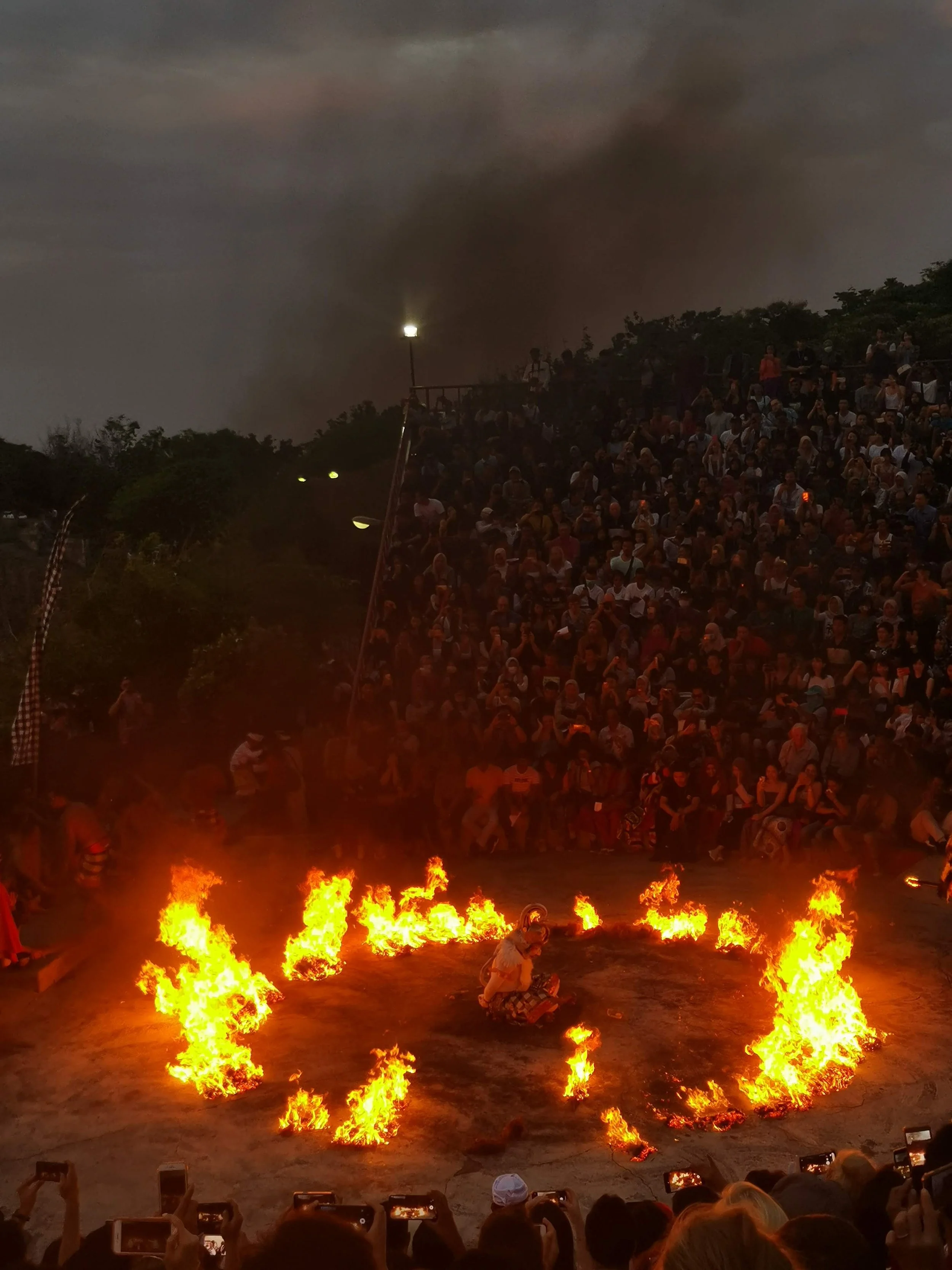A large crowd of people watching a fire dance performance outdoors at night, with flames forming a circle around a performer sitting in the center.