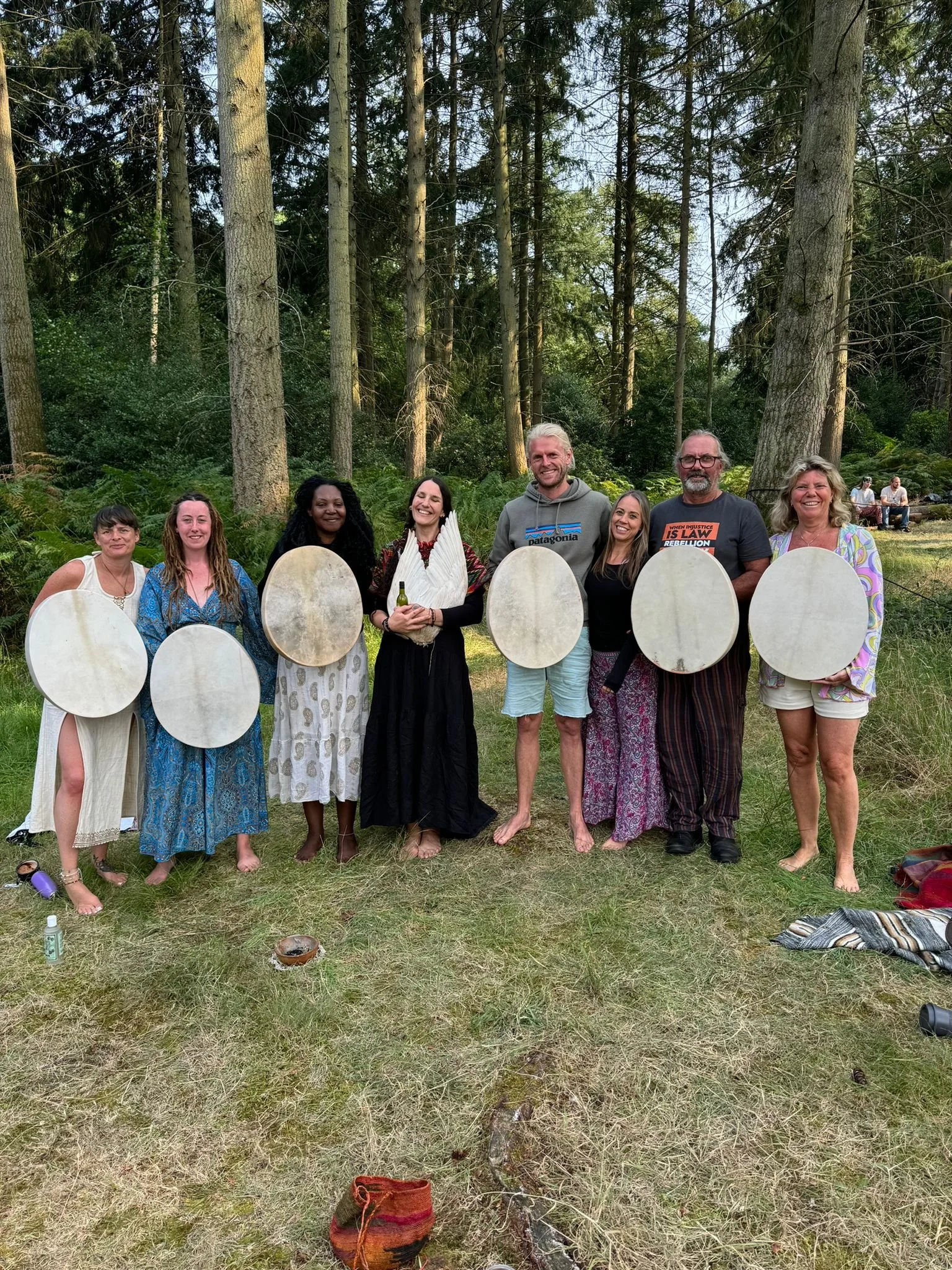 Group of eight people standing outdoors in a forest, holding large circular white shields, with some individuals smiling and dressed in casual and bohemian clothing.
