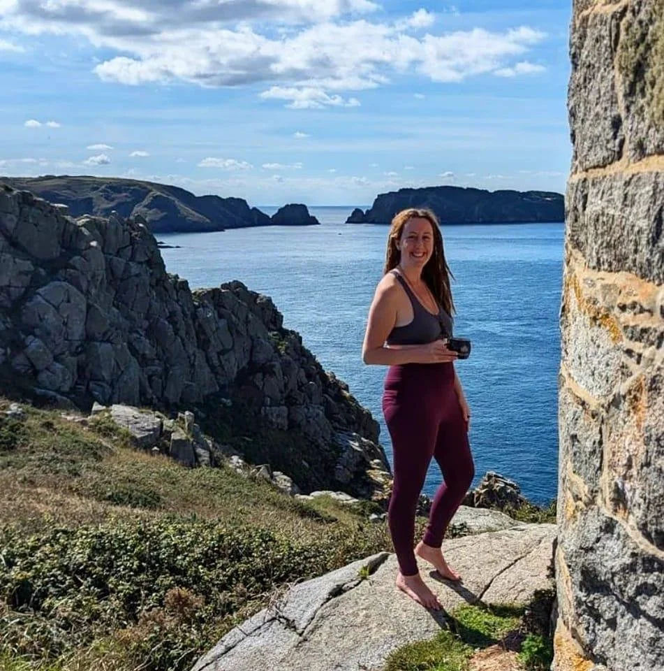 A woman standing barefoot on rocky terrain near the coast, holding a camera, with a scenic view of cliffs, the ocean, and sky in the background.