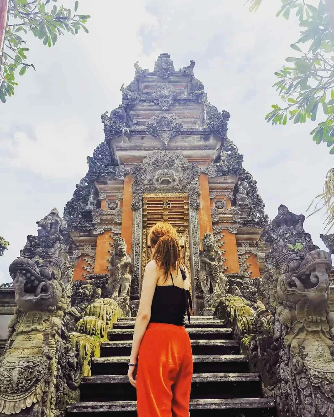 Woman with reddish hair, wearing a black top and orange pants, standing on stone steps leading to an ornate, ancient temple with intricate carvings and stone statues.