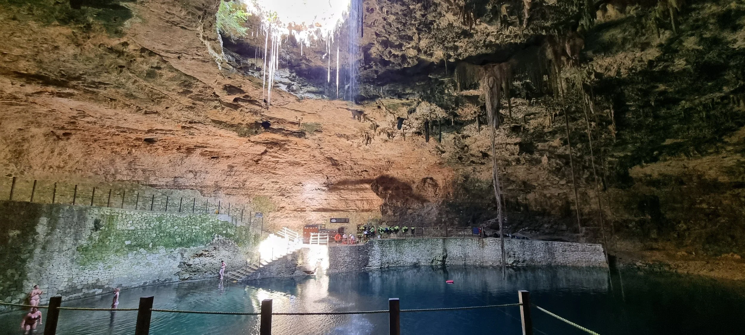 People swimming in a pool inside a large cave with stalactites hanging from the ceiling and a large opening allowing sunlight to enter.