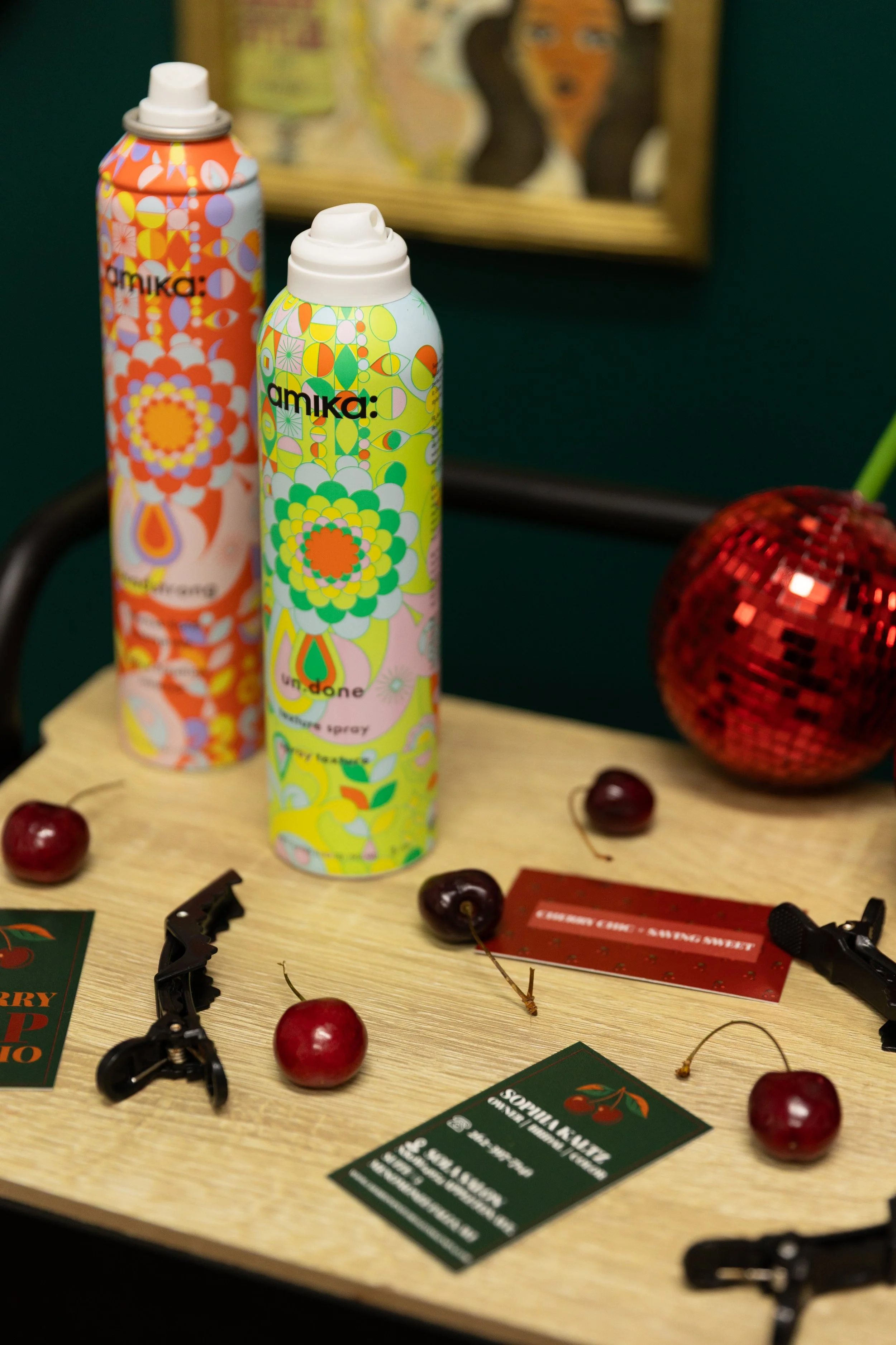 Two cans of Amika dry texture spray with colorful, floral patterns on a wooden surface, accompanied by cherries, hair clips, a festive red sequined ball, and a small red gift card, with a framed artwork in the background.