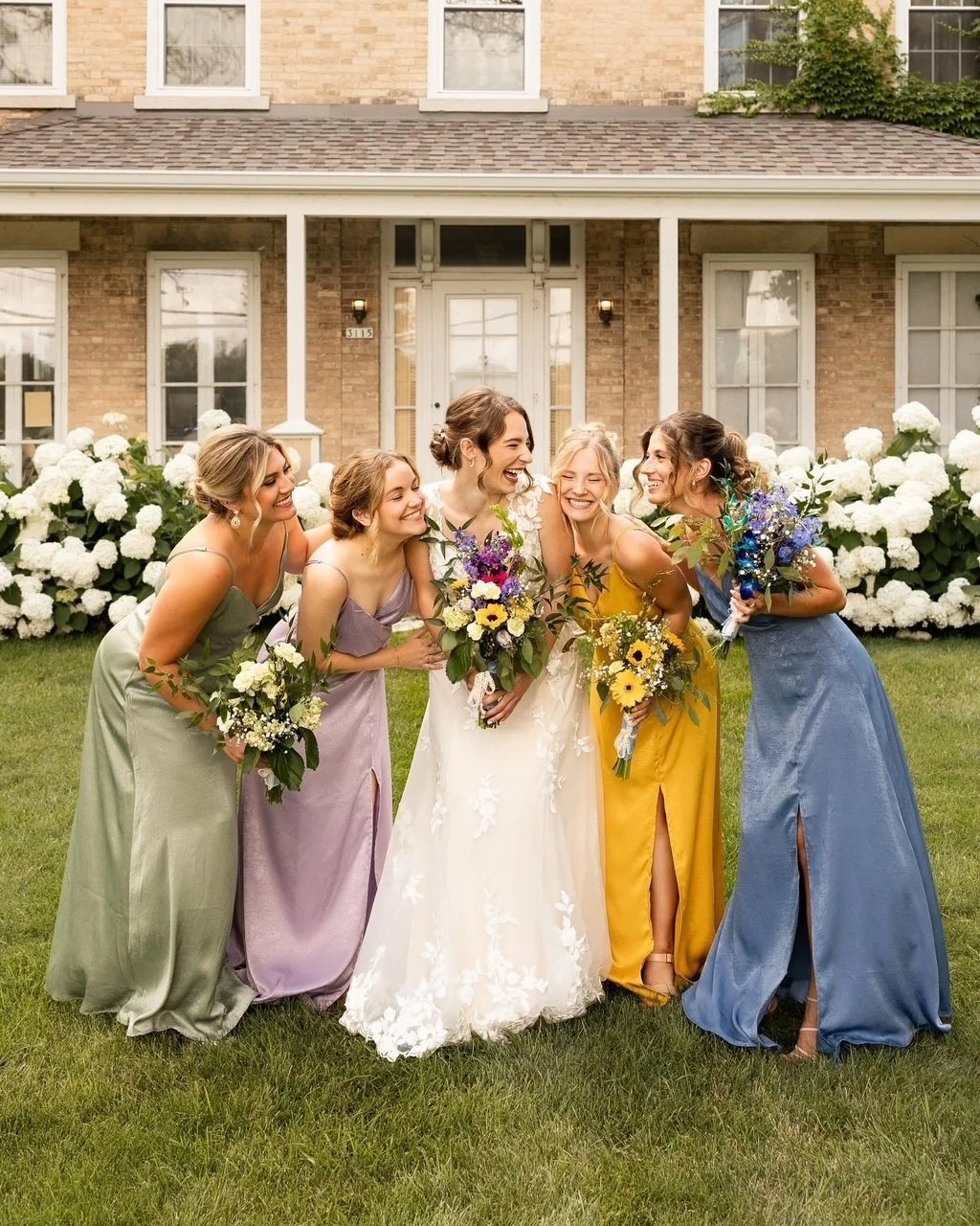 Bride and five bridesmaids smiling and holding bouquets in front of a house with white flowers.