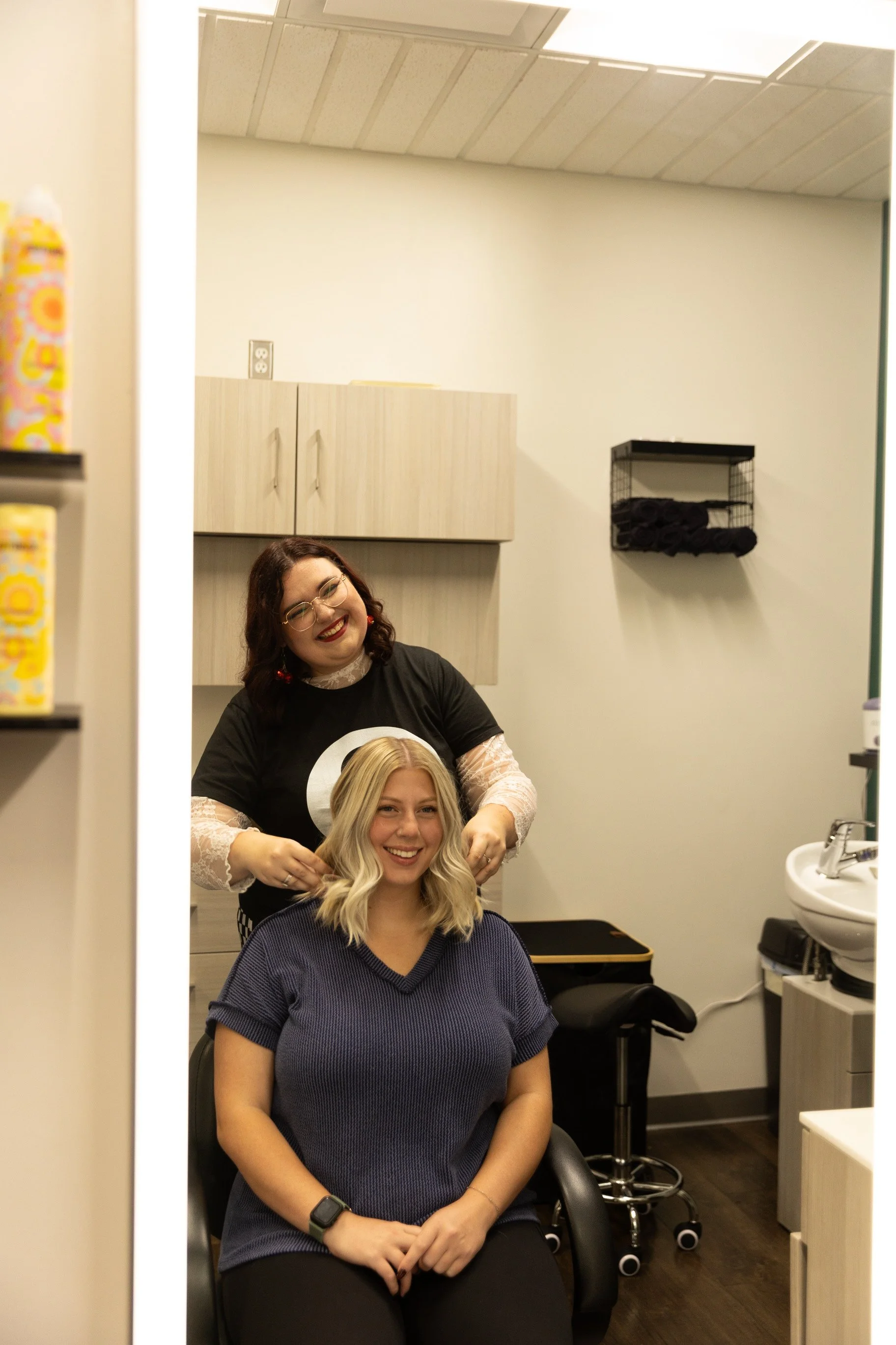 A woman with blonde hair sitting in a salon chair getting her hair styled by a woman with glasses and brunette hair in a hair salon.