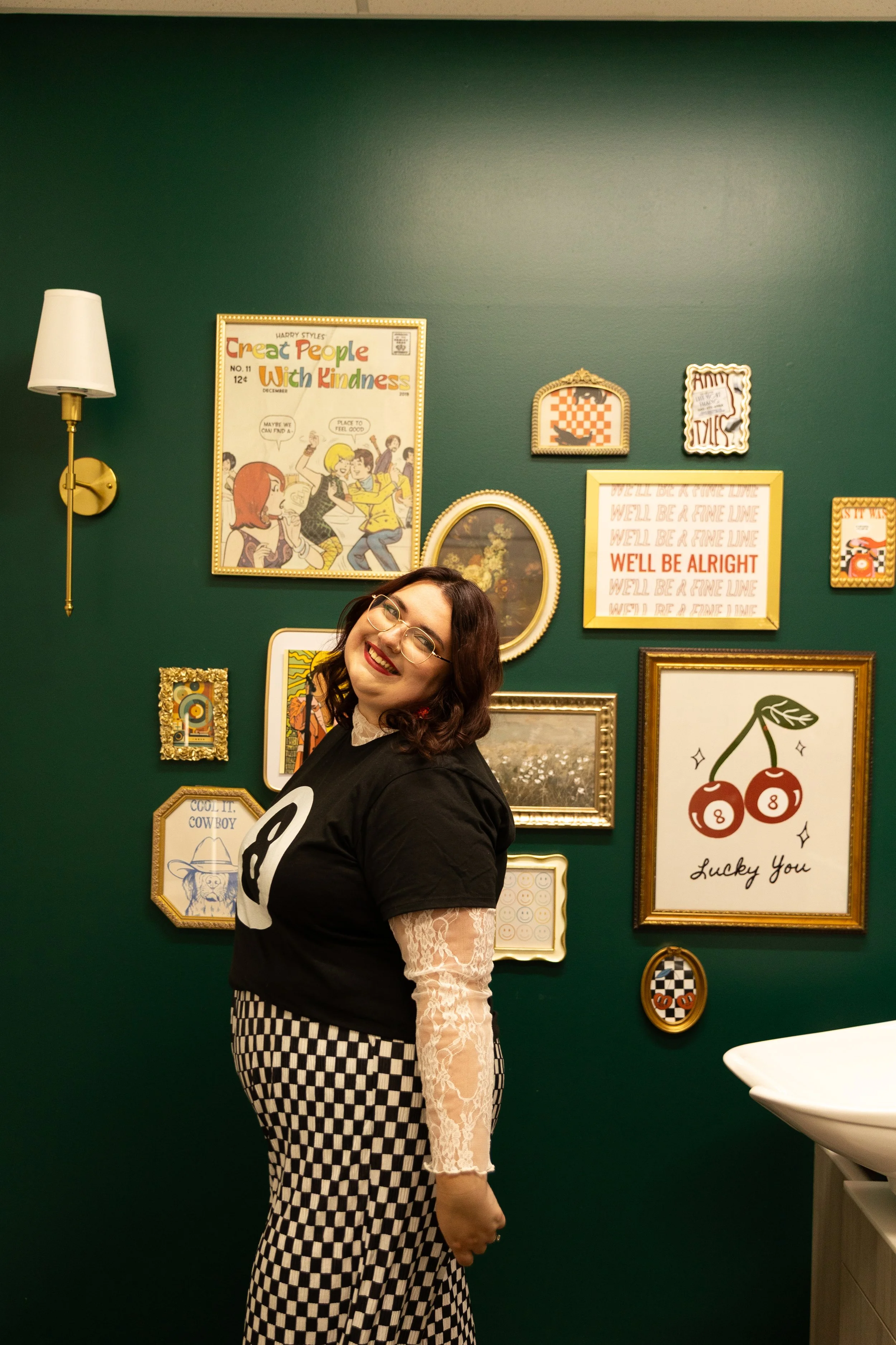 A smiling woman with glasses and red lipstick wearing a black shirt with a skull graphic, checkered pants, and a lace sleeve shirt standing in front of a green wall decorated with various framed pictures and artwork.