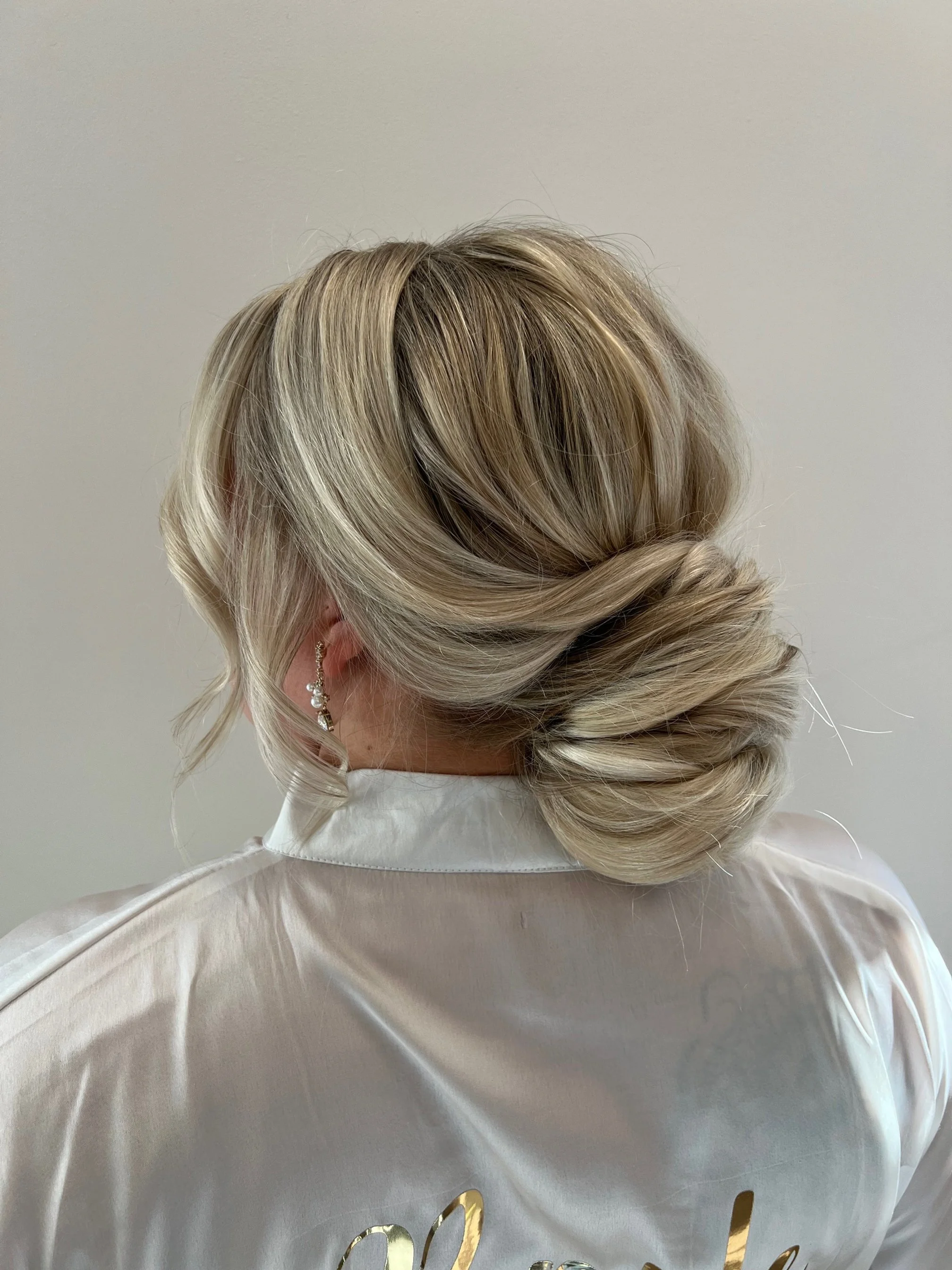 Back view of a woman with blonde hair styled in an elegant low updo, wearing a white satin robe with gold lettering and a dangling earring.