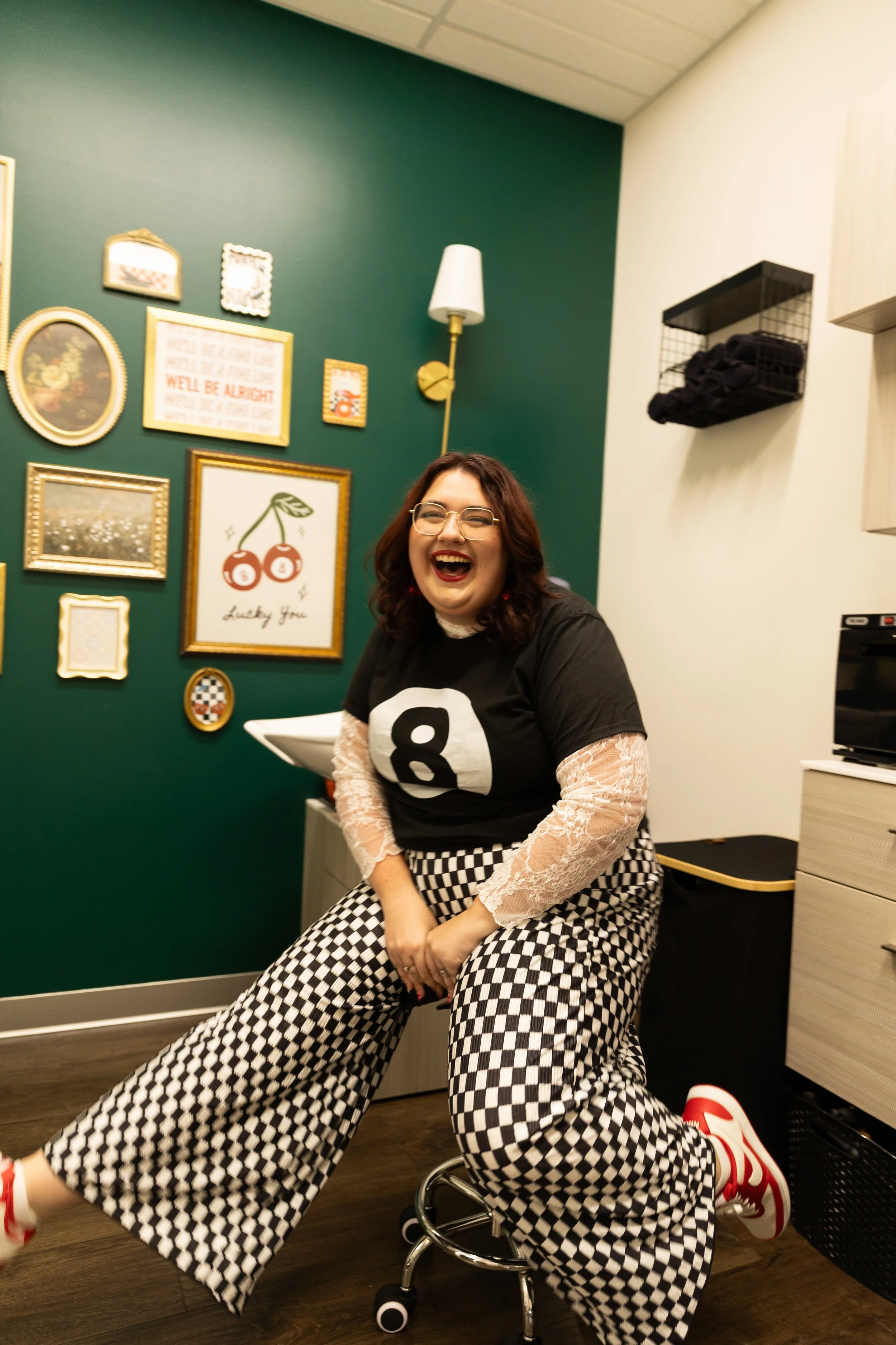 A woman with glasses and dark hair laughing, sitting on a stool in a room with a green accented wall decorated with framed pictures and signs. She wears a black t-shirt with a white number 8, checkered wide-leg pants, and white and red sneakers.