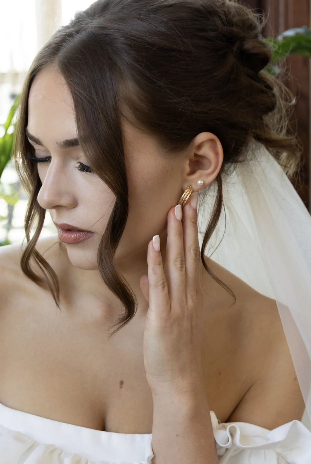 Close-up of a woman with brown hair styled in loose curls, wearing pearl and gold earrings, and a white off-shoulder dress, touching her ear with her hand, with a bridal veil on her shoulder, indoors with natural light and green plants in the background.