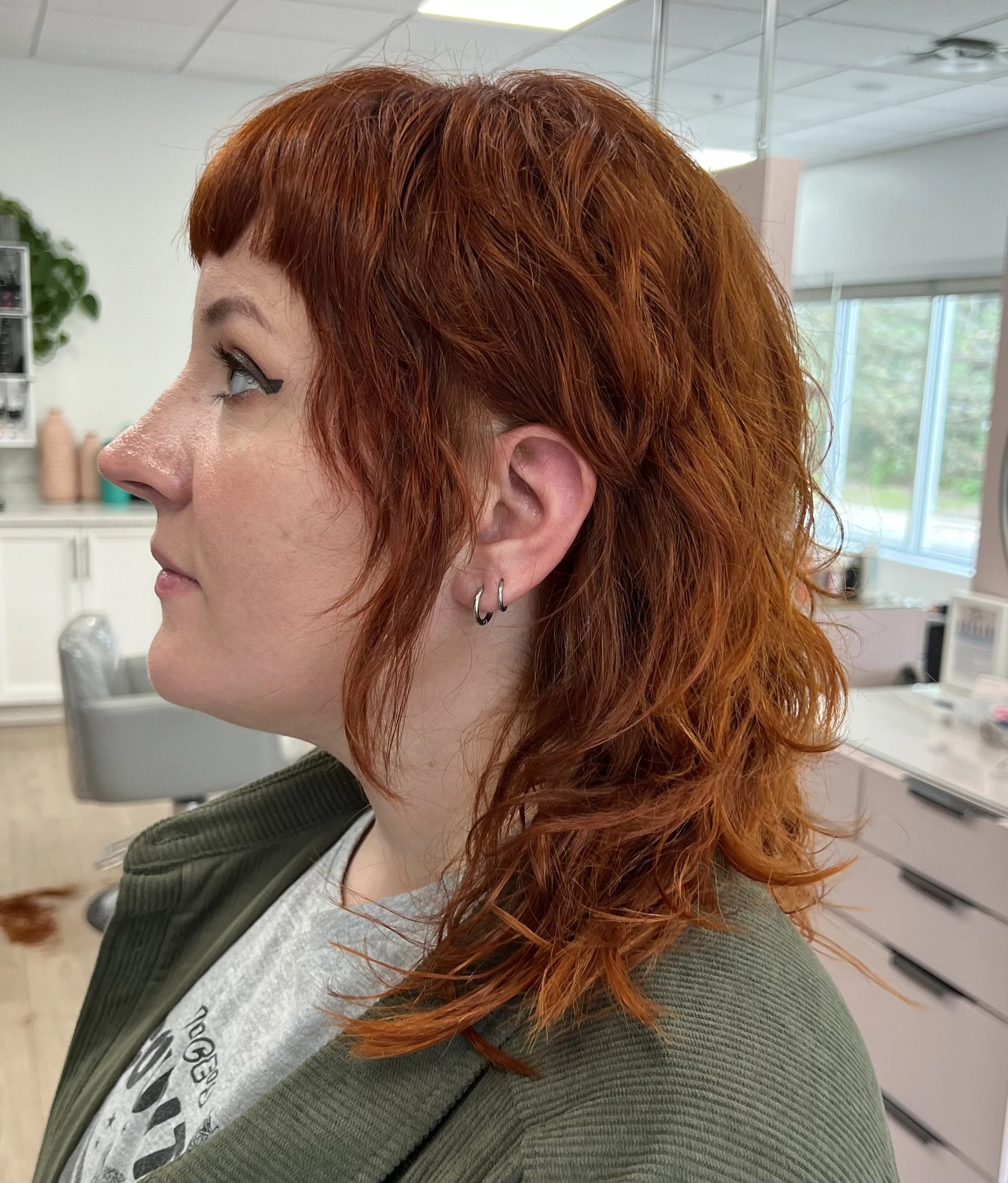 Side profile of a woman with curly, shoulder-length red hair, wearing earrings and a gray shirt with a green jacket, in a hair salon.