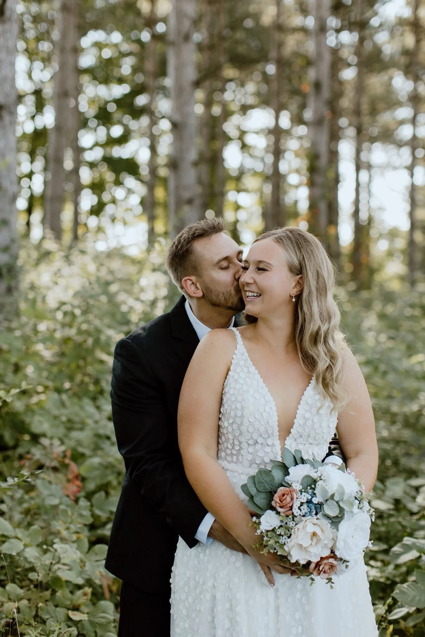 A newlywed couple standing outdoors in a forest. The groom, dressed in a black suit, is kissing the bride on the cheek. The bride, with wavy blonde hair, is wearing a white wedding dress with a deep V neckline and holding a bouquet of white, peach, and blue flowers.
