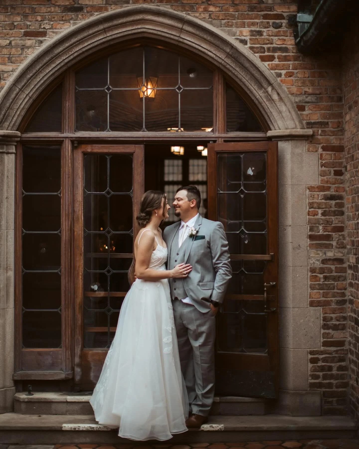 Bride and groom sharing a romantic moment in front of an arched wooden and glass door, inside a brick building, at their wedding.