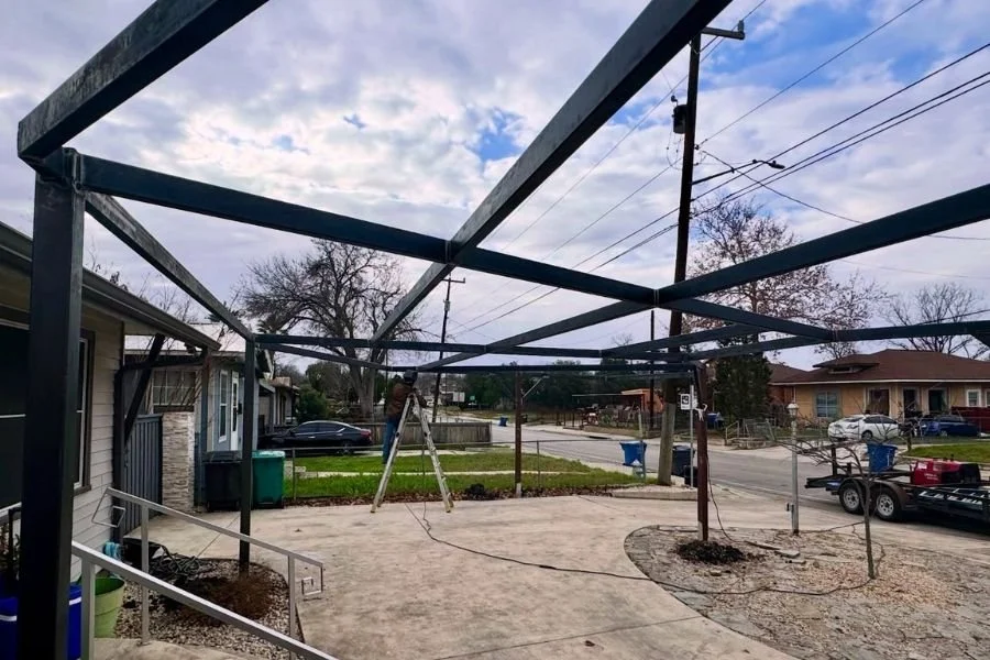 Steel frame structure under construction for a carport or patio cover, showing exposed beams and posts set over a residential driveway.