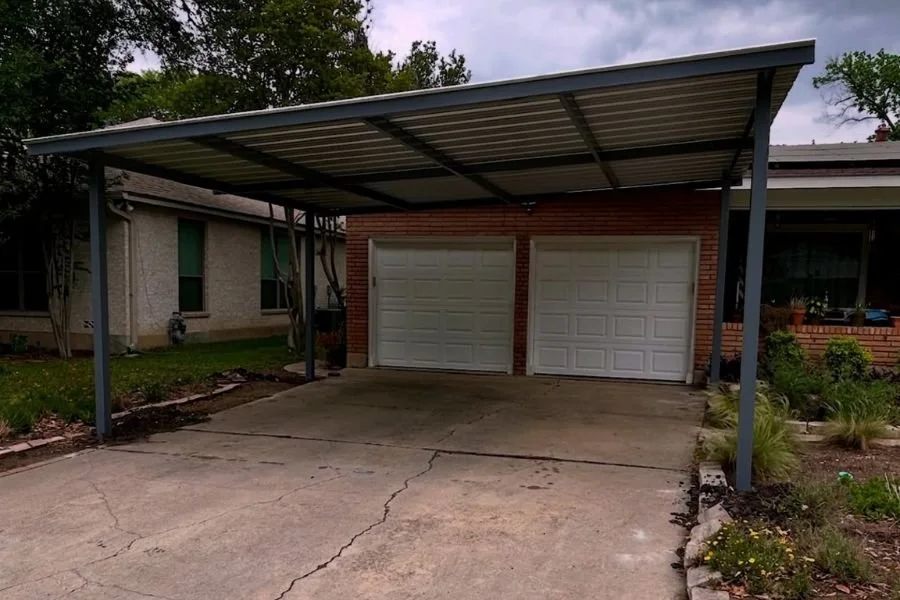 Front view of a double garage with a large metal carport and patio cover extension, providing covered parking over a cracked concrete driveway