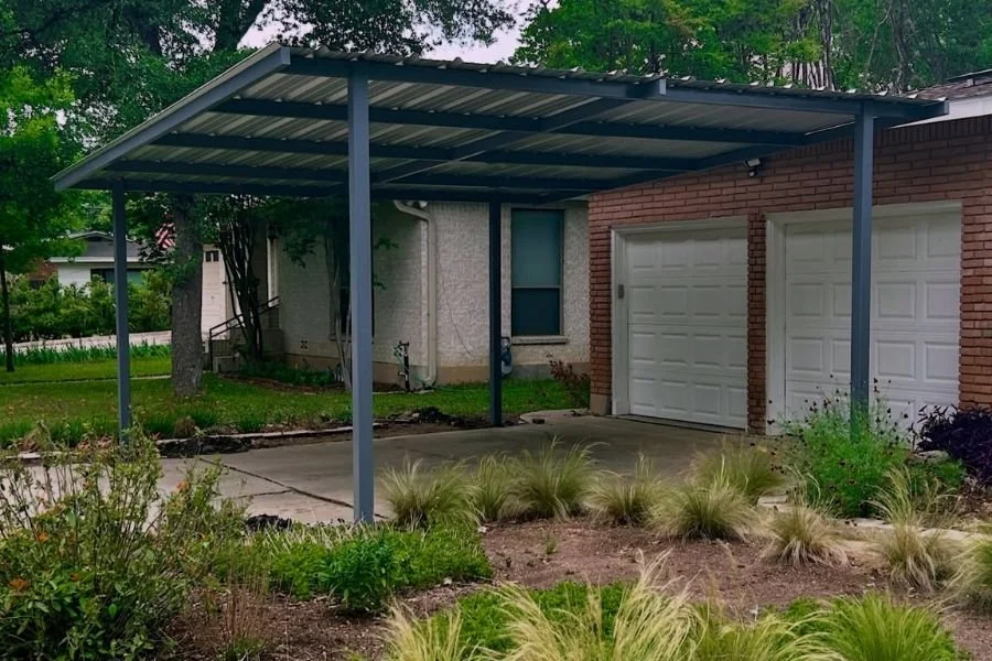 Modern metal carport attached to a brick home, featuring a sloped steel roof supported by gray posts over a concrete driveway.