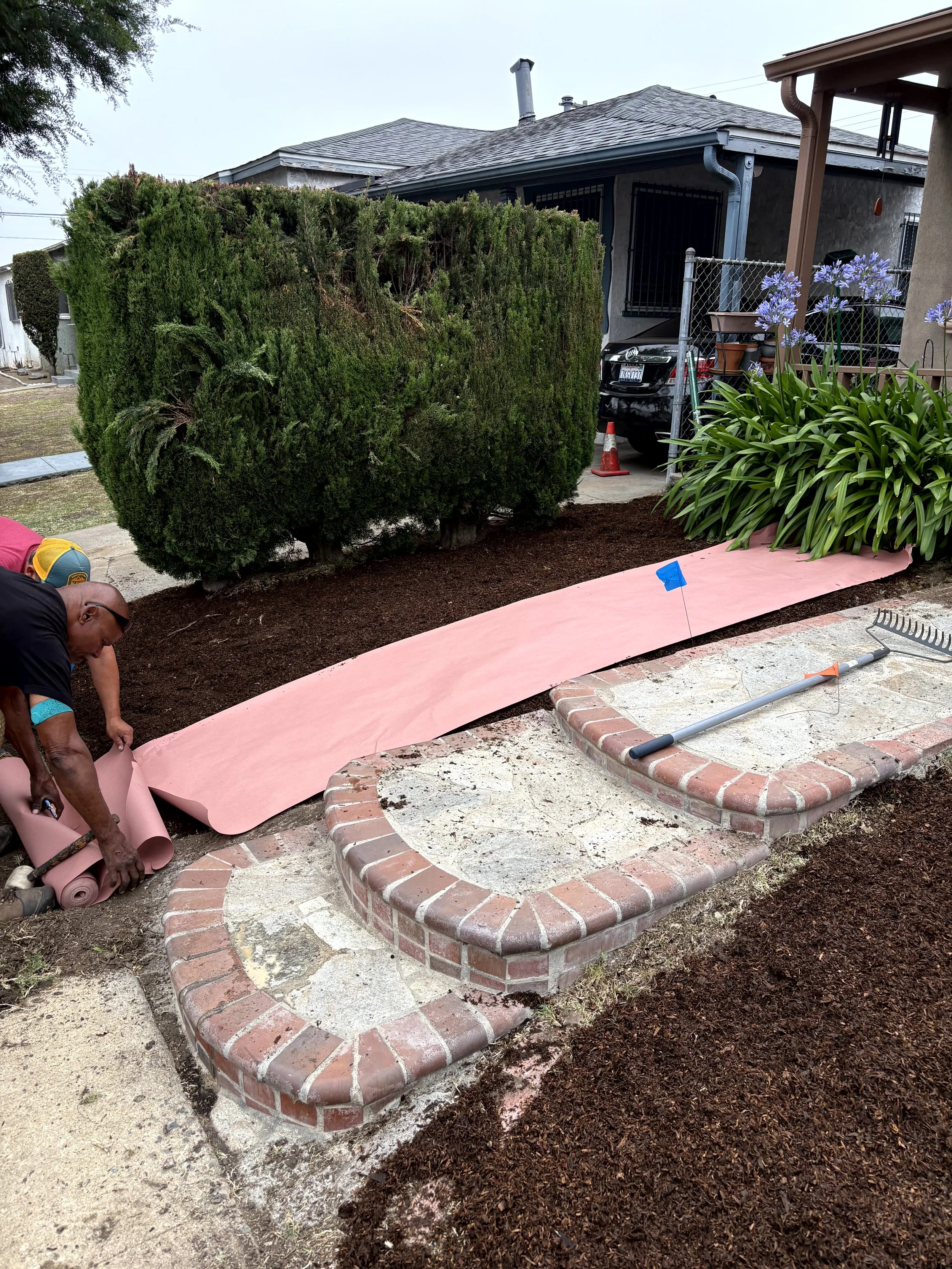 People preparing a garden bed with a brick border and composted soil in a residential yard, with a large bush, plants, a house, a car, and gardening tools visible.