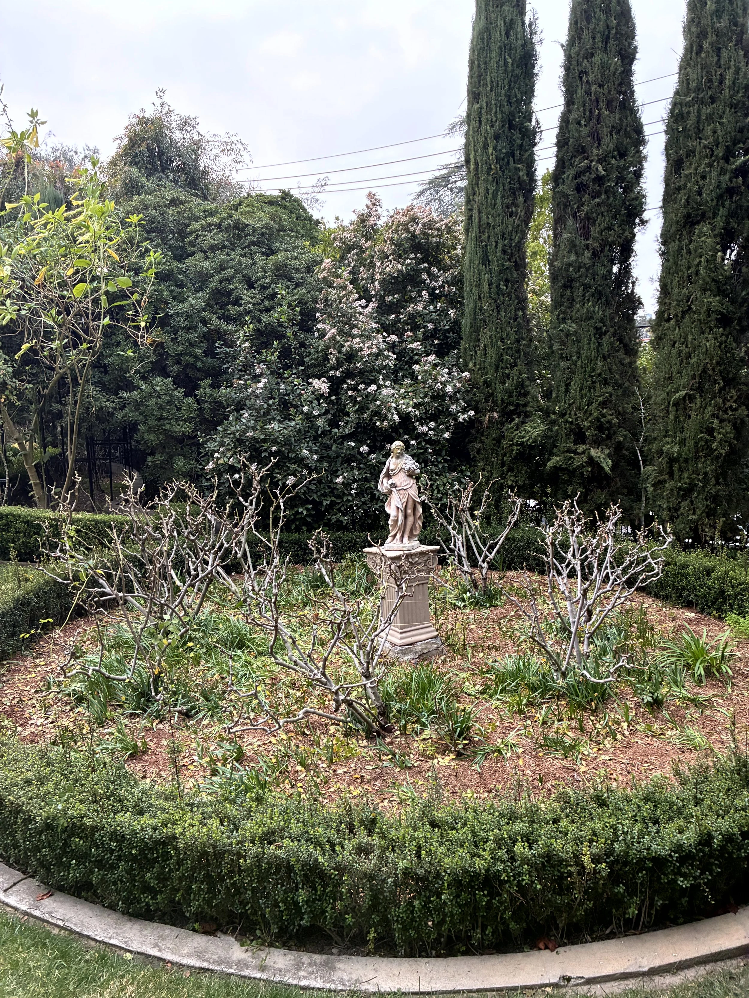A garden with a statue of a woman holding a flower, surrounded by various green bushes and tall trees, with a cloudy sky overhead.