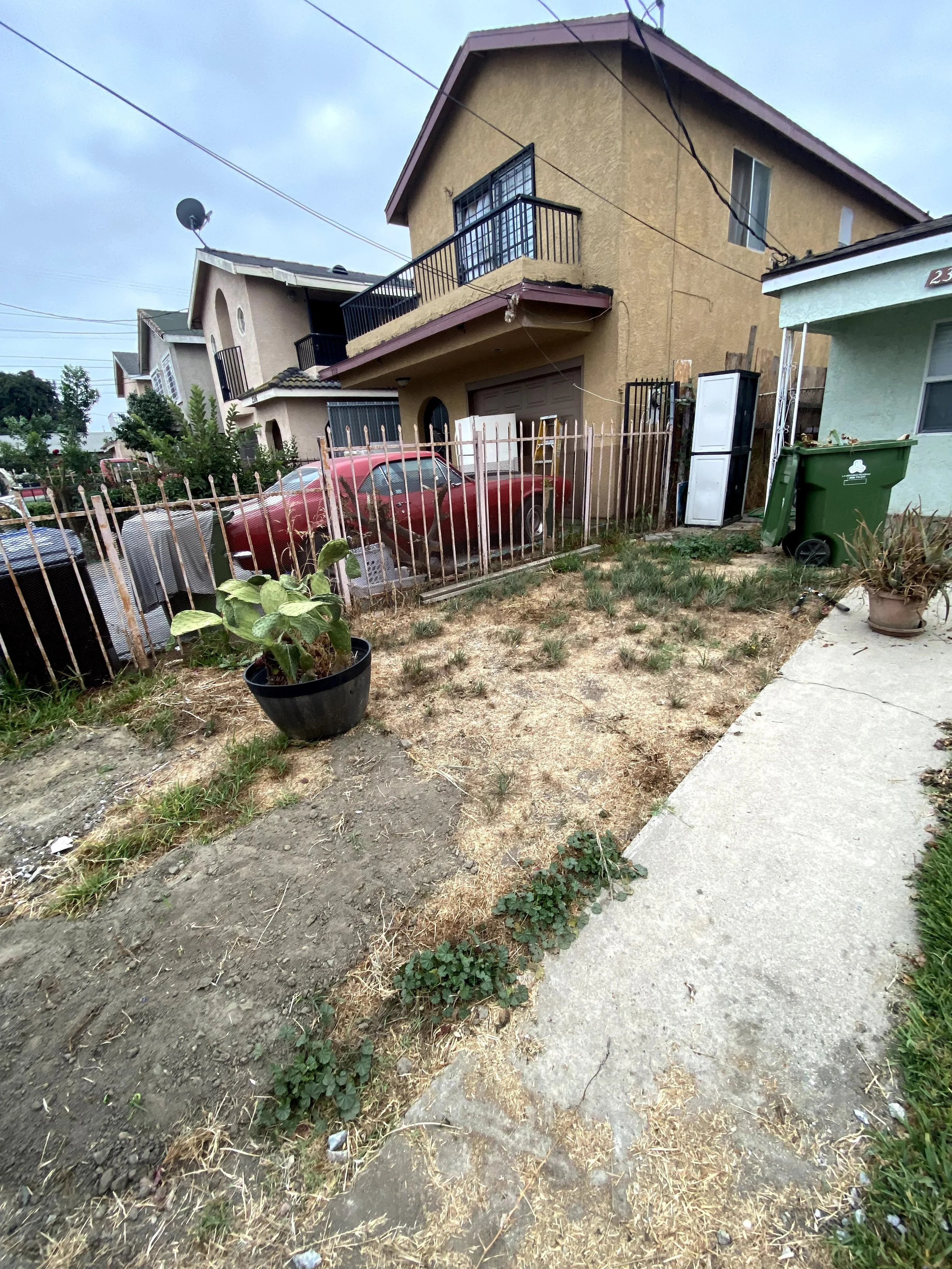 A two-story yellow house with a small balcony and black metal railing, situated behind a black metal fence. In the front yard, there is a red vintage car parked behind the fence, with a green trash bin and a white cabinet outside the house. The yard has dry grass, patches of dirt, and some potted plants near the sidewalk. Overhead power lines are visible against a cloudy sky.