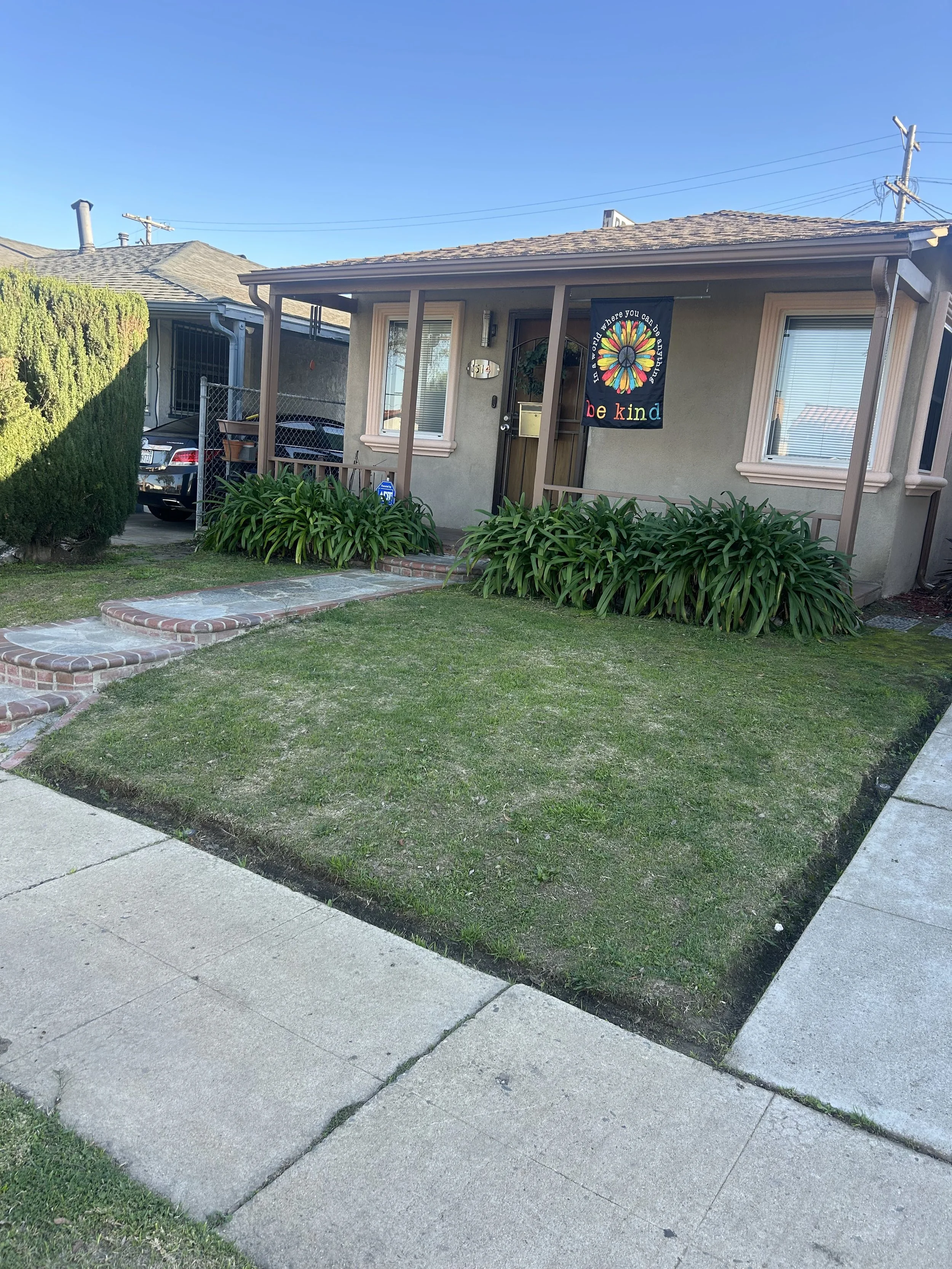 Front yard of a house with a well-maintained grass lawn, concrete sidewalk, and plants near the porch. The house has beige walls, pink window frames, and a door with a wreath. There's a banner hanging on the porch that reads, 'See how bright you can be when you be kind.' A black car is parked in the driveway surrounded by a chain-link fence. The sky is clear and blue.