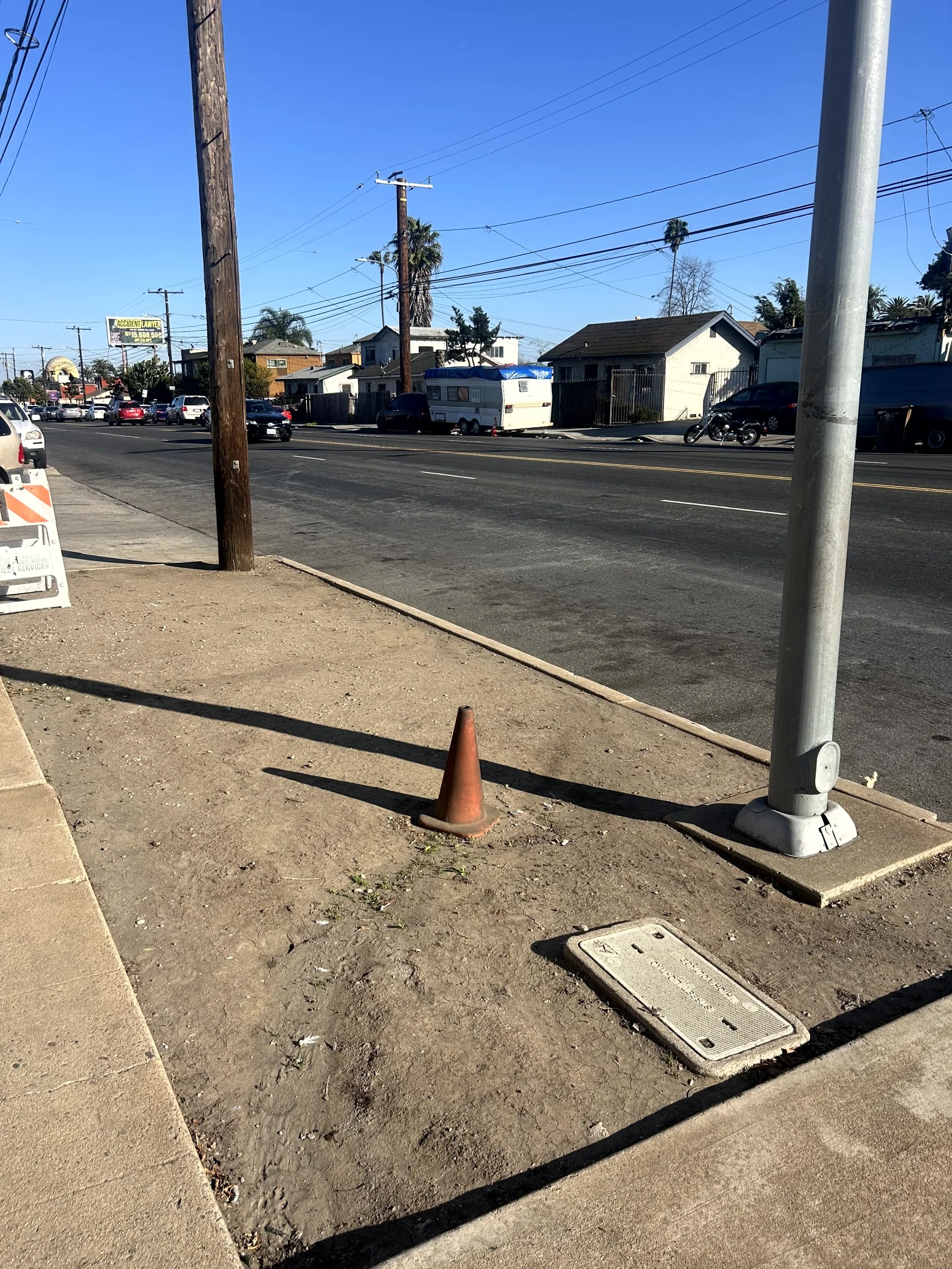 Sidewalk with an orange traffic cone, manhole cover, and utility pole with attached light pole. Street with parked cars and houses in the background under clear blue sky.