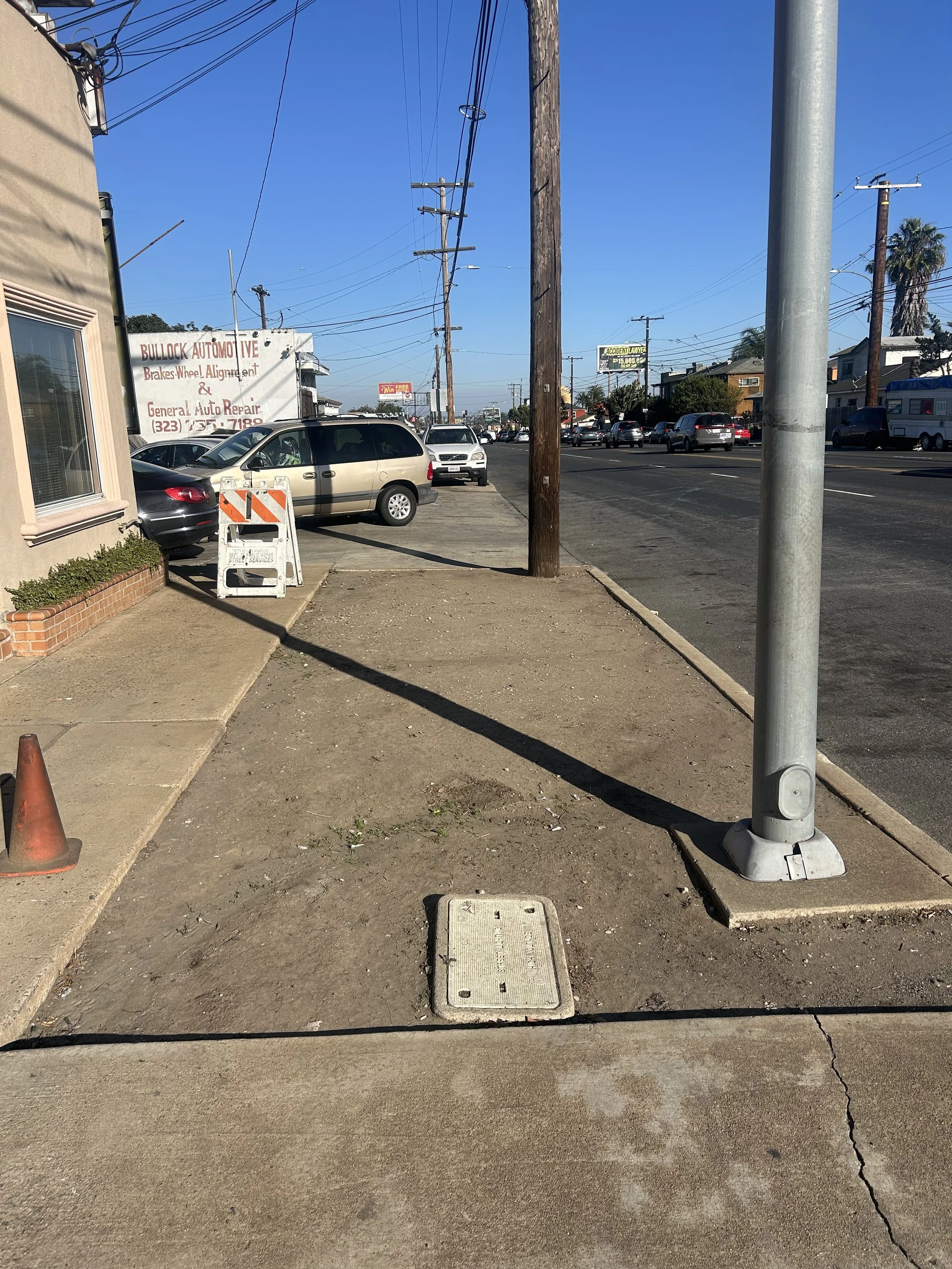 Sidewalk with construction sign, utility pole, manhole cover, and parked cars on a busy street under a clear blue sky.