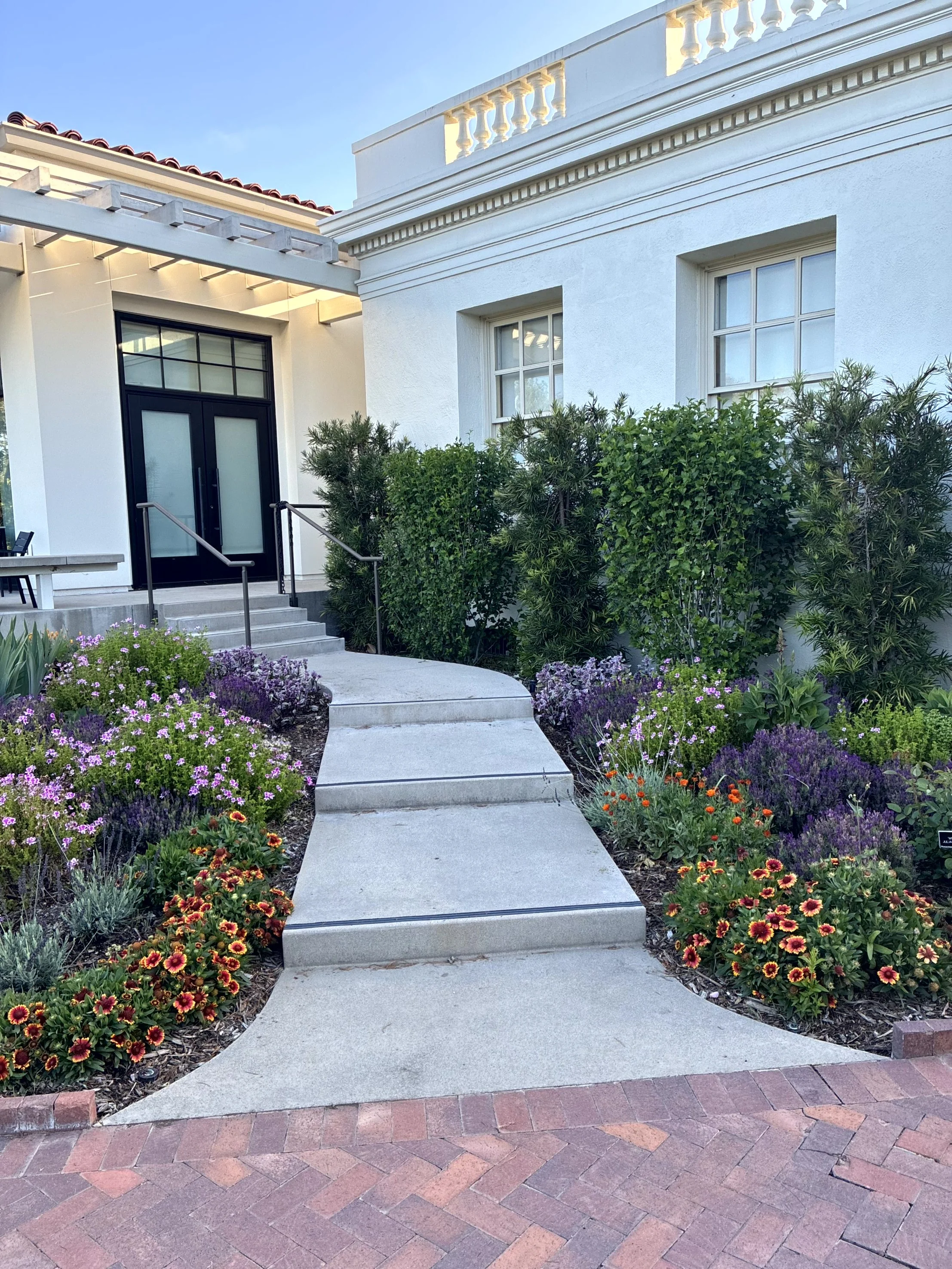White house with two windows, a decorative balcony ledge, and a black front door with glass panes. A concrete walkway with stairs and black handrails leads up to the door. The house is surrounded by lush, colorful flower beds and green shrubs, with a clear blue sky overhead.