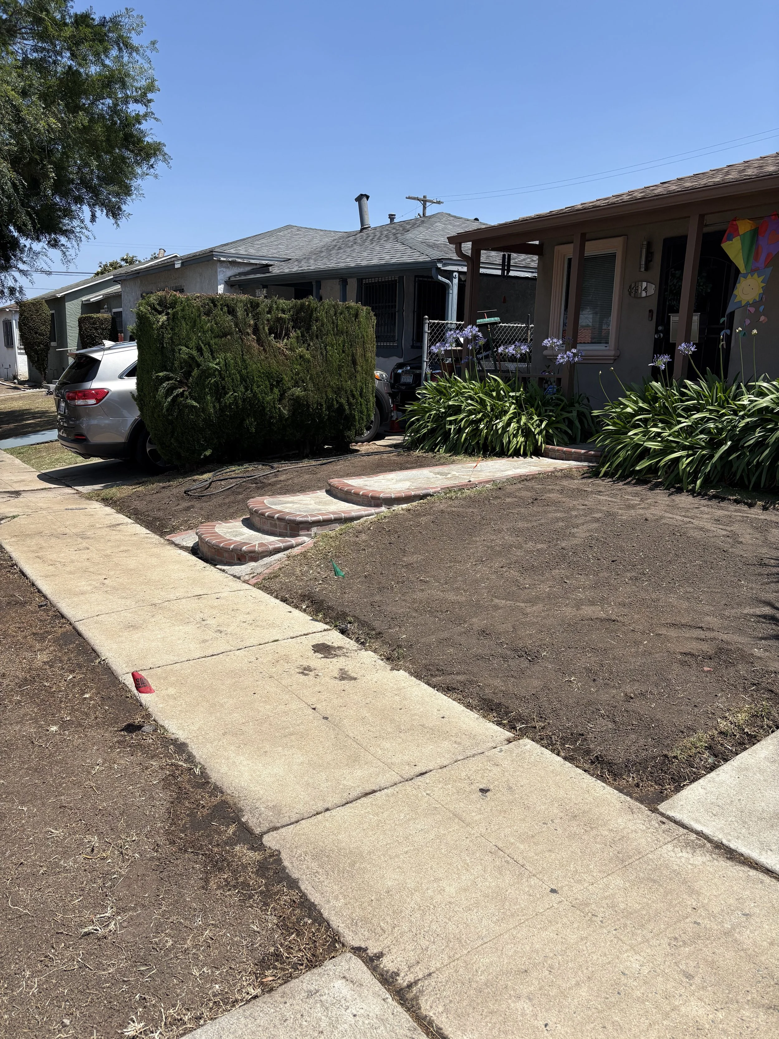 Residential front yard with a sidewalk, a bush covering part of a parked silver SUV, garden with purple flowers, and a house with a porch and a kite decoration