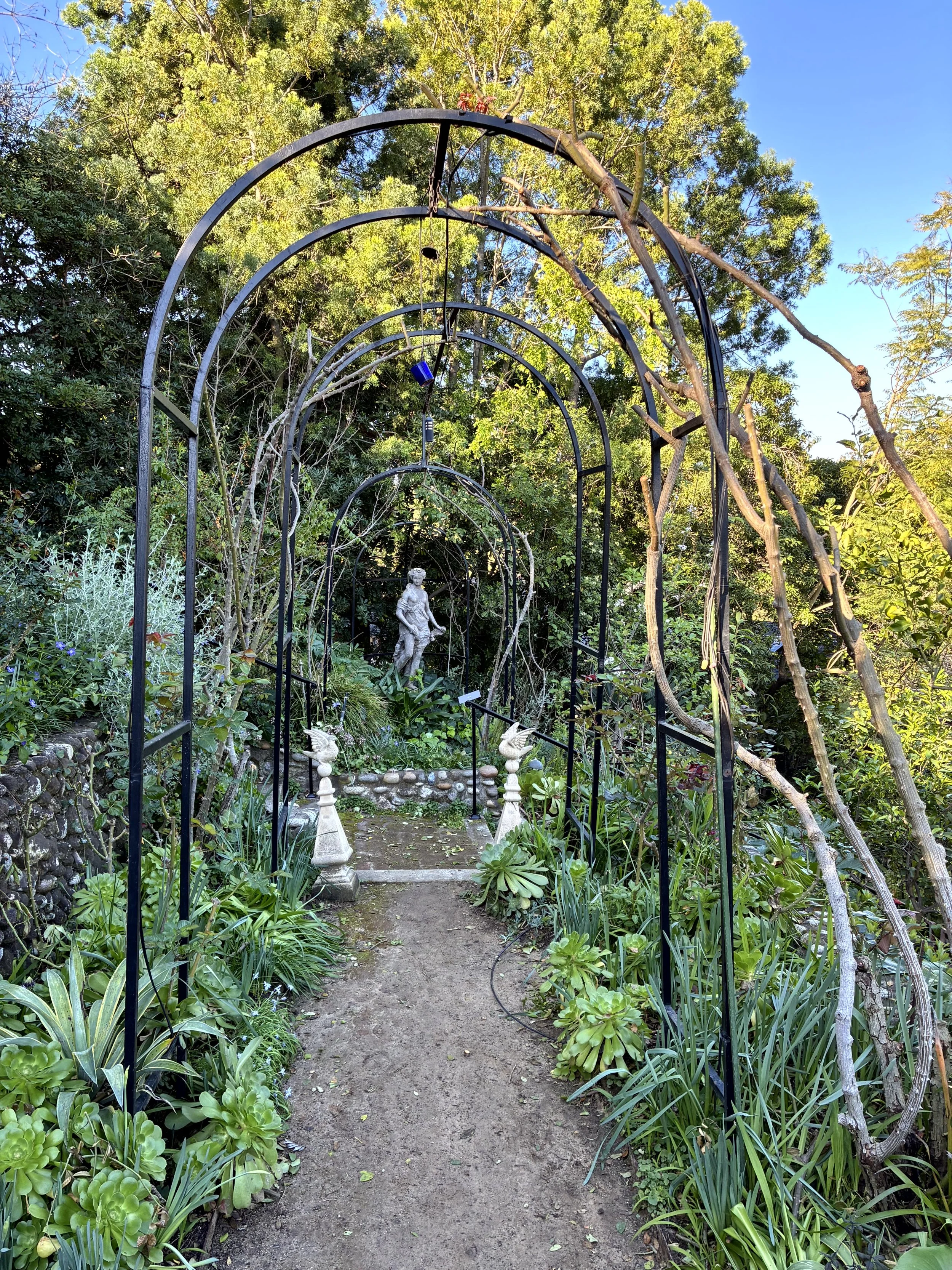 A garden pathway surrounded by lush green plants and trees, with a black metal garden archway leading to a statue of a girl, and some garden ornaments on either side of the path.