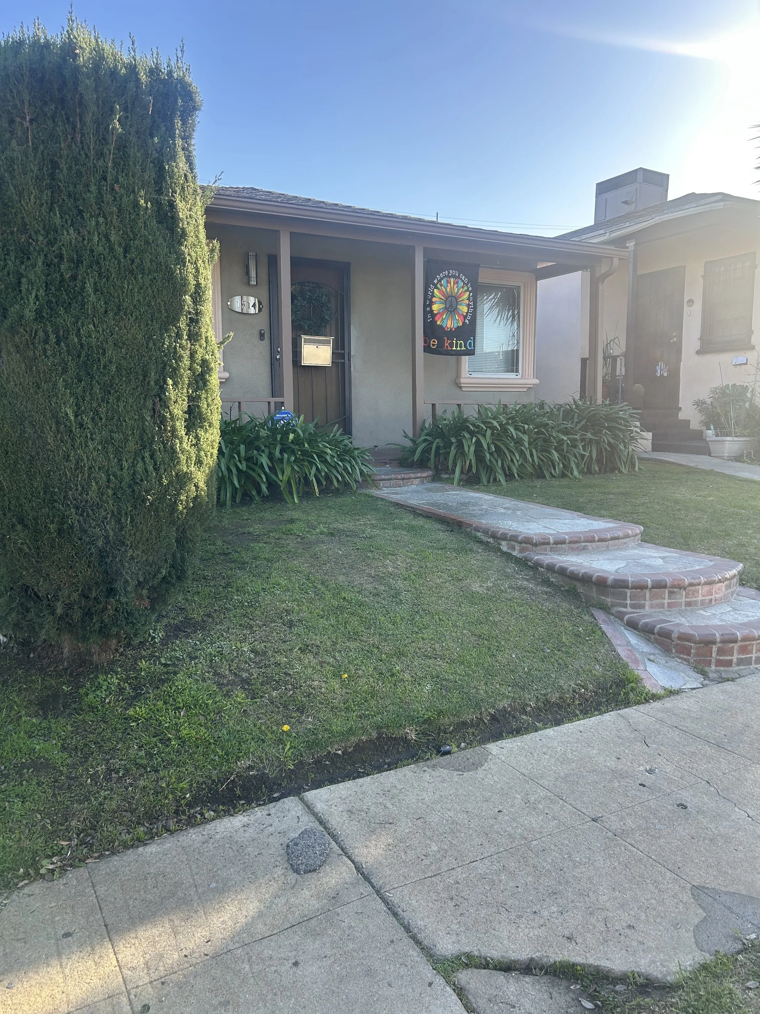 A residential front yard with a sidewalk, a green lawn, a large tree on the left, and a small front porch with steps, plants, and a black door with a colorful sign.
