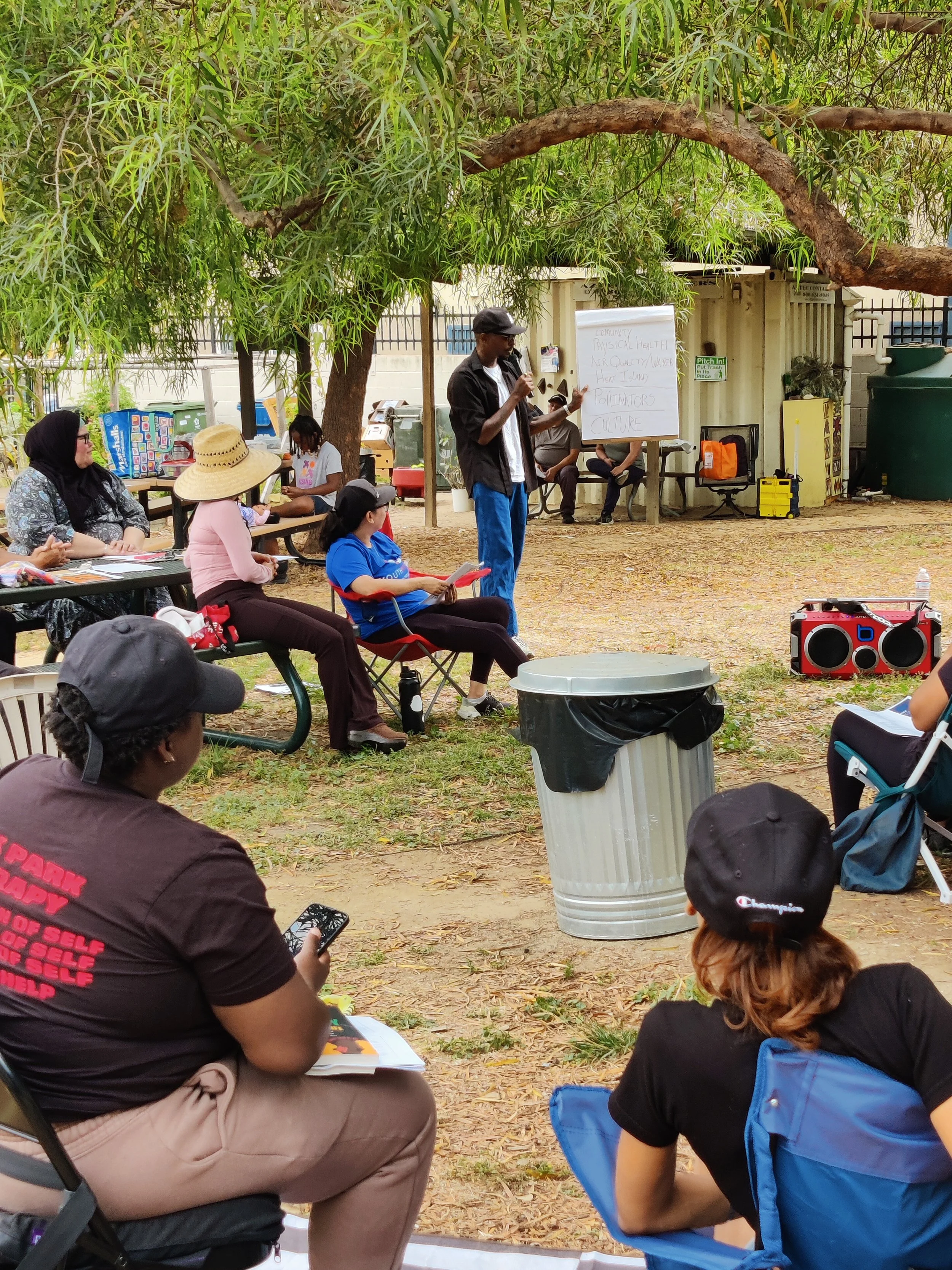 A man is speaking to a group outdoors under a large tree, with a whiteboard displaying writing about community issues such as health, air quality, and culture. Several people are seated and listening, some taking notes or using electronic devices.