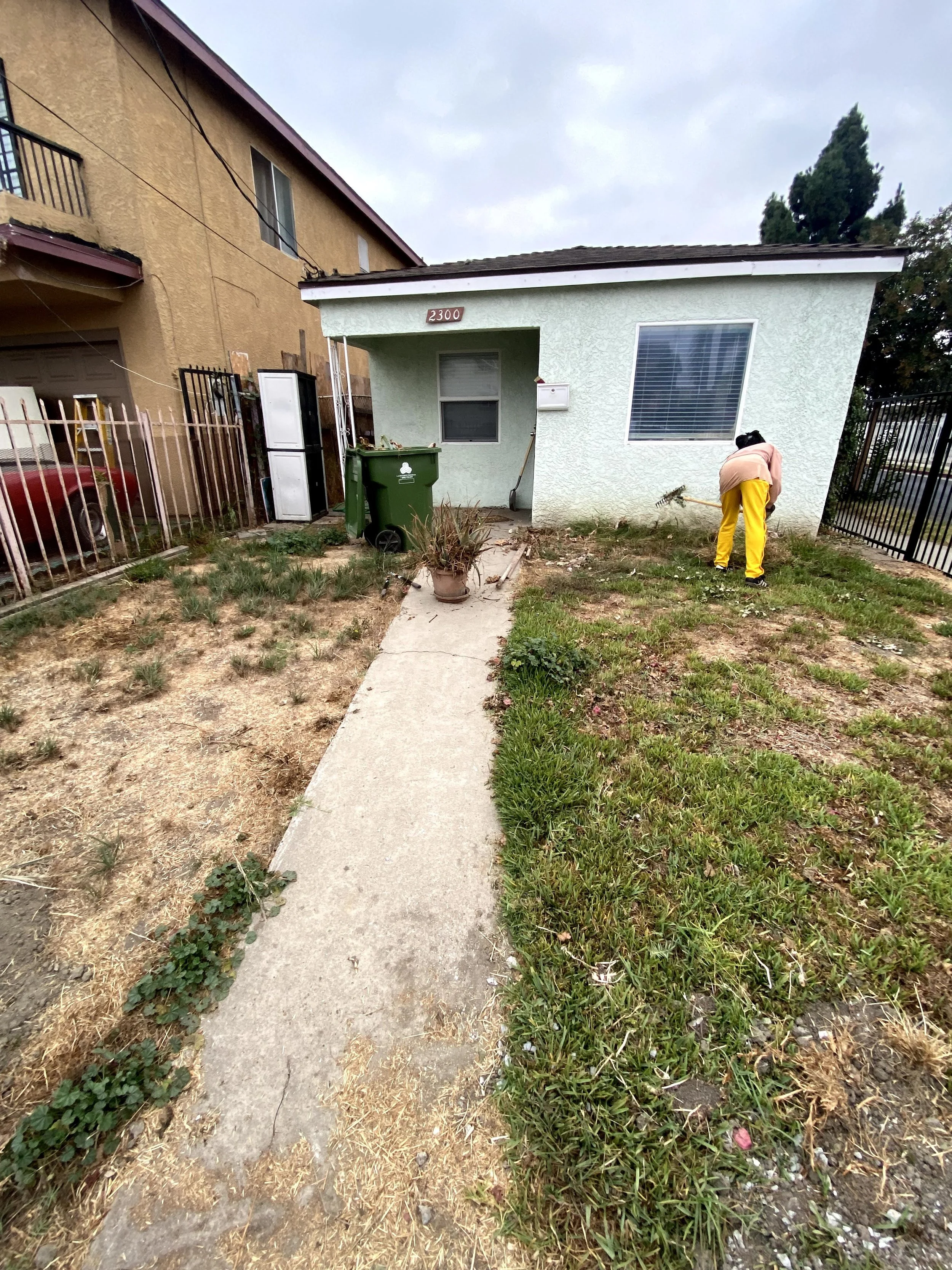 Person in yellow pants and light jacket raking leaves in front yard of a house with light green stucco walls and a slanted roof, with a house number 2300 above the front porch.