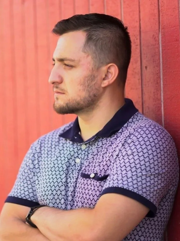 A young man with short dark hair and a beard, standing against a red wooden wall, looking to the left with arms crossed, wearing a patterned short-sleeve polo shirt.