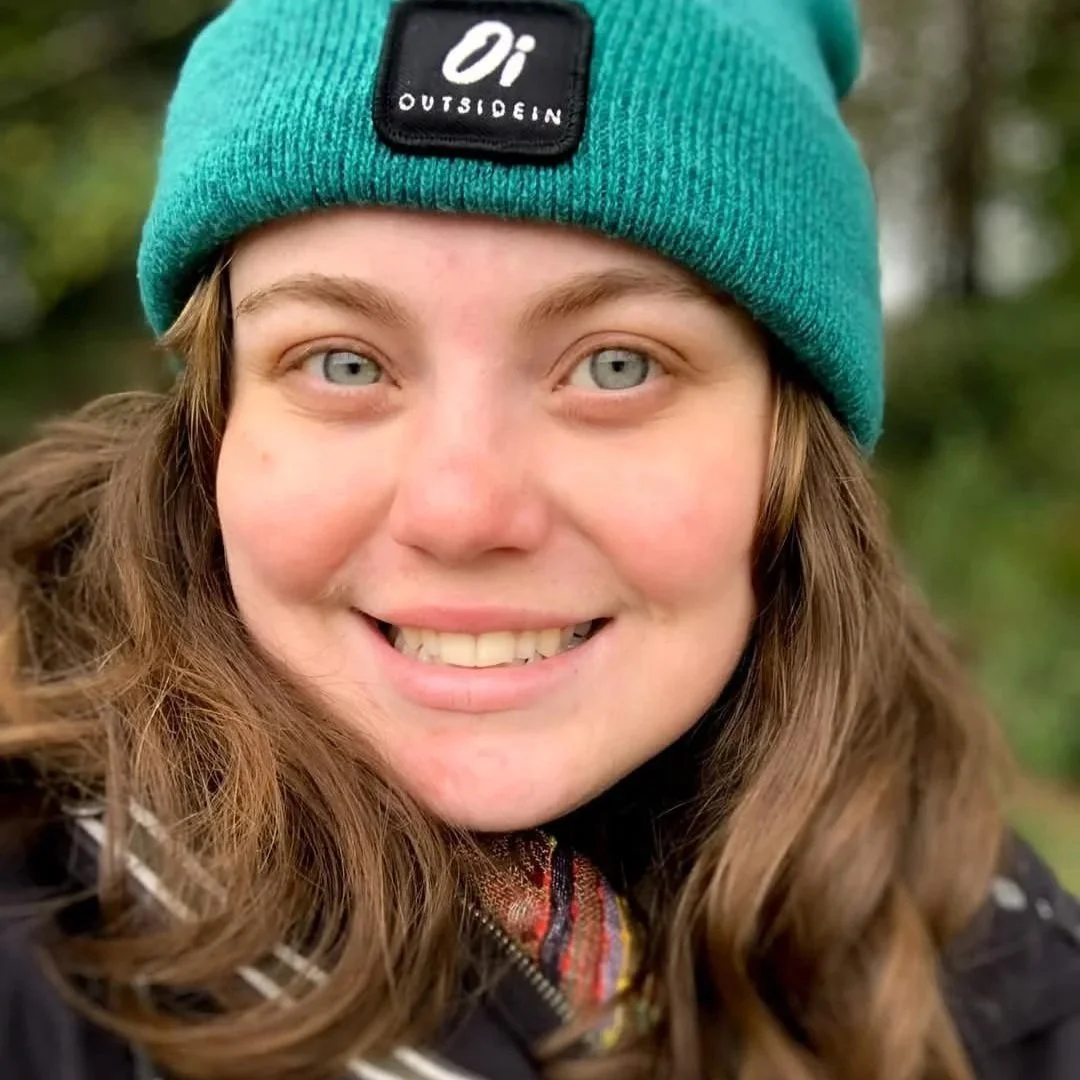 Close-up of a smiling woman with long brown hair wearing a teal knit beanie with the label "Oi OUTSIDEIN".