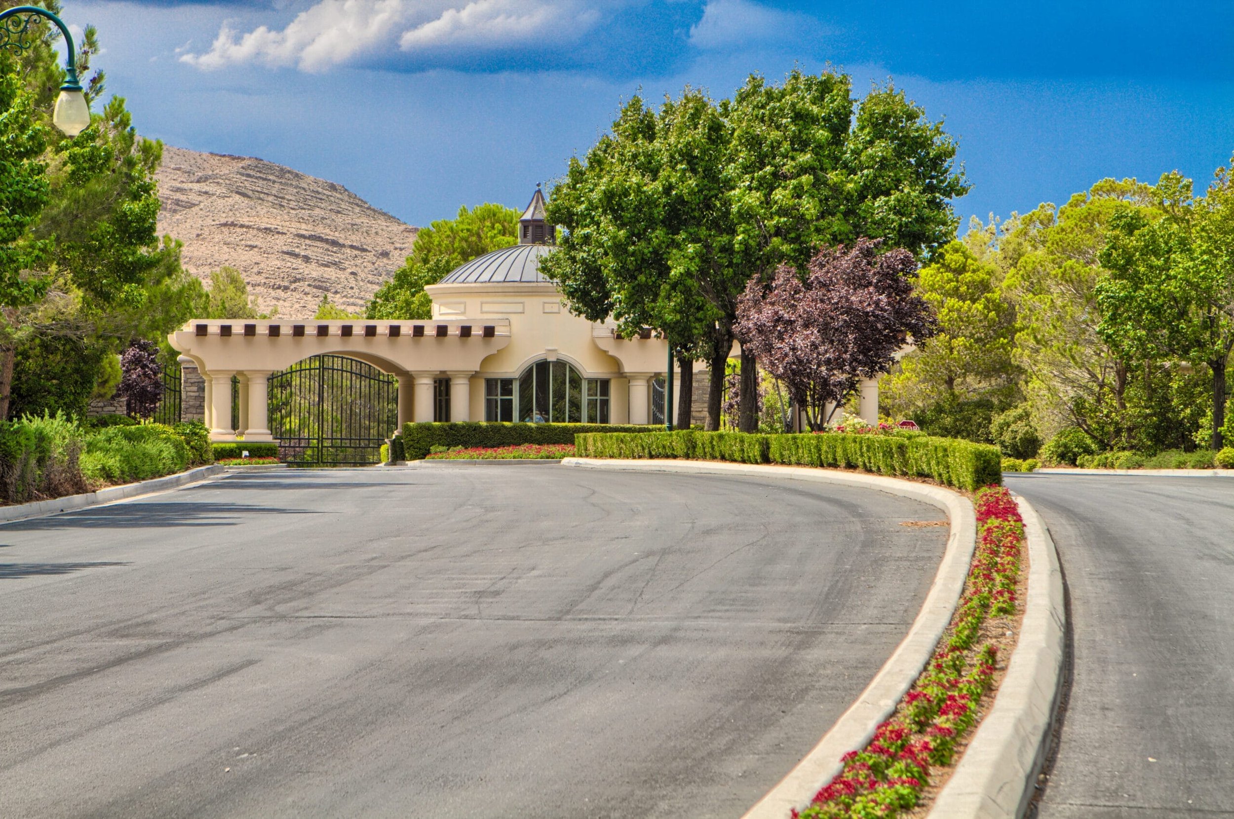 Gated entrance to Southern Highlands Golf Club, with a curved driveway, lush green trees, colorful flower beds, and a building with a dome-shaped roof in the background under a partly cloudy sky.
