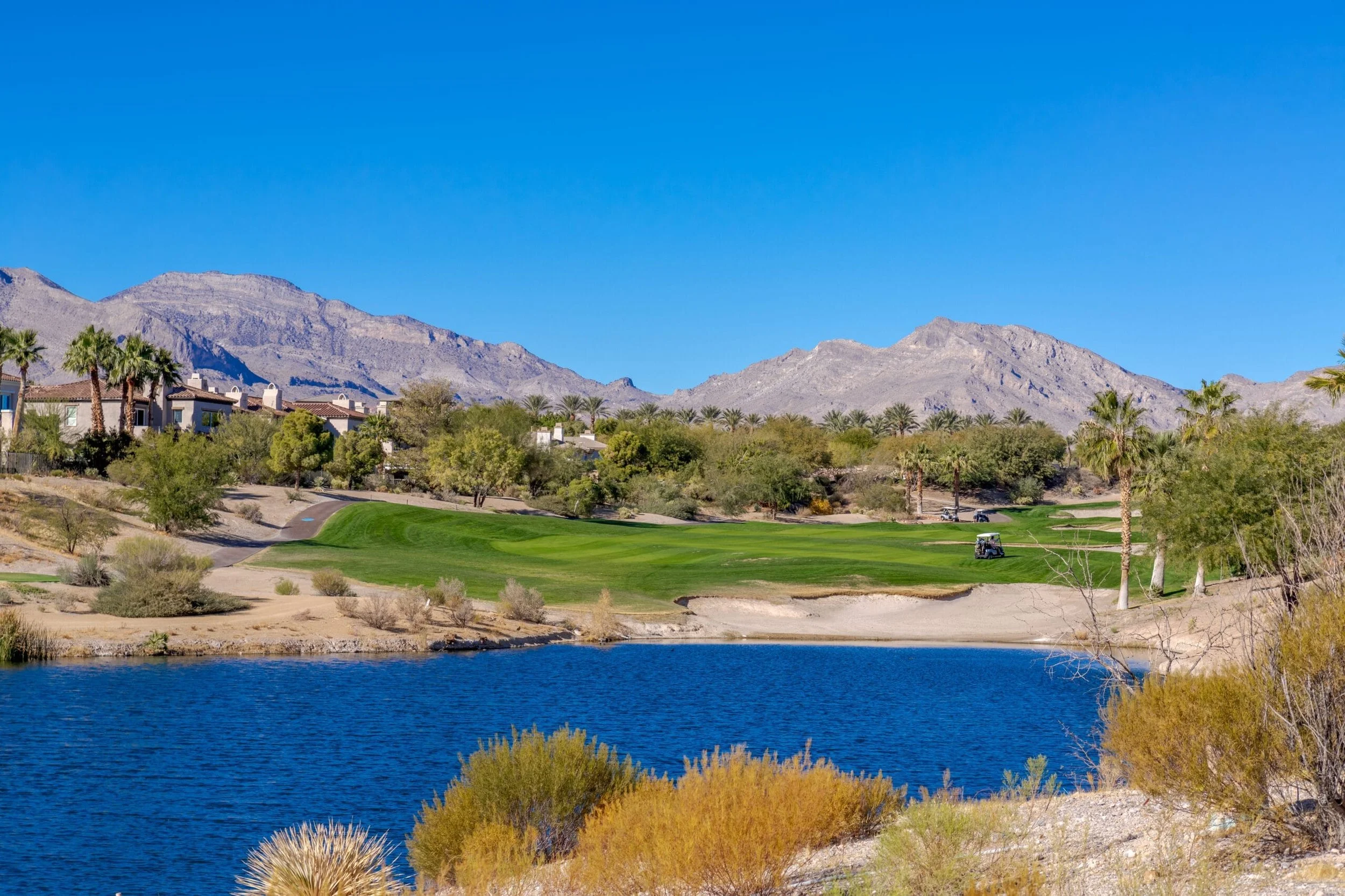Scenic view of a golf course with a water hazard, surrounded by desert vegetation and residential buildings, with mountains in the background and a clear blue sky.