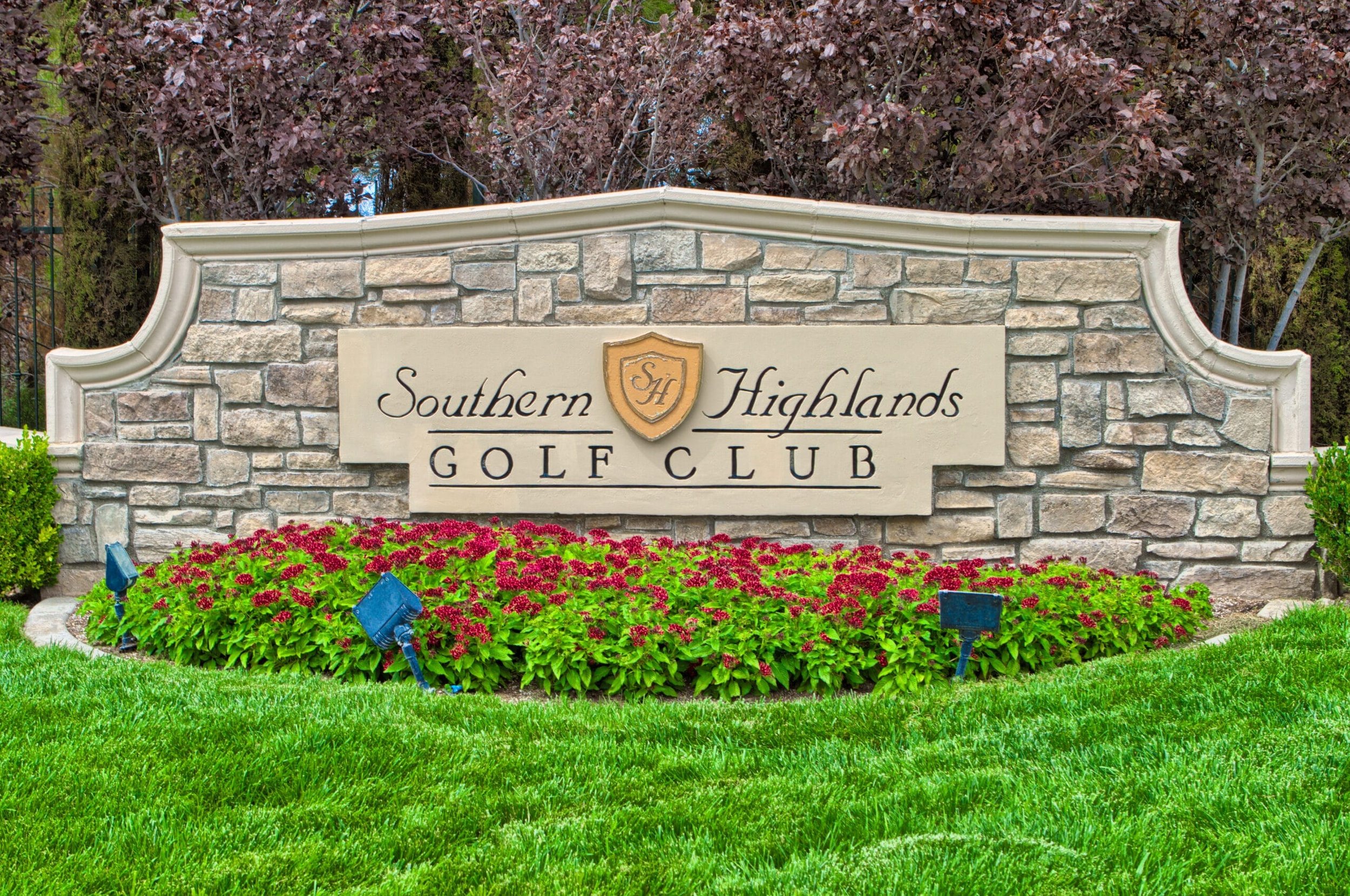 Entrance sign for Southern Highlands Golf Club with a stone background, a crest, and a flower bed with pink and green plants in front.
