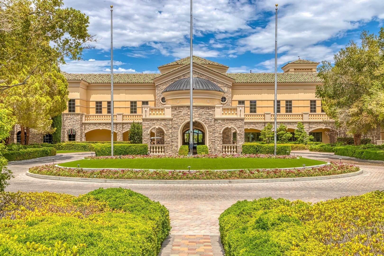 A large, elegant estate in Southern Highlands Golf Club features a stone and stucco facade, a covered entrance arch, manicured lawns, vibrant flower beds, trees, and flagpoles in a sunny setting under a partly cloudy sky.