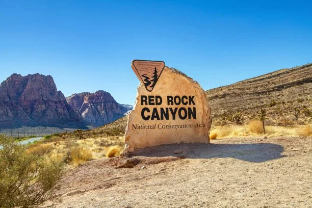 Sign for Red Rock Canyon in a desert landscape with mountains in the background and a clear blue sky.