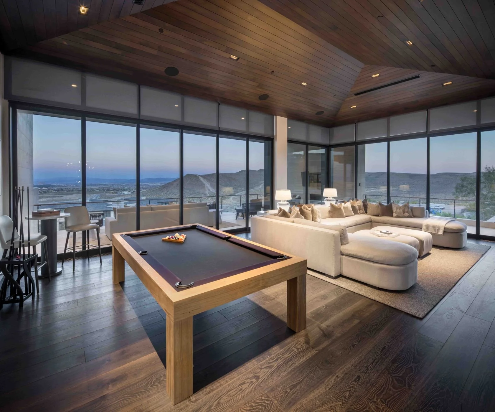 Modern living room with large windows, a pool table, and a sectional sofa, overlooking mountains and a distant landscape.
