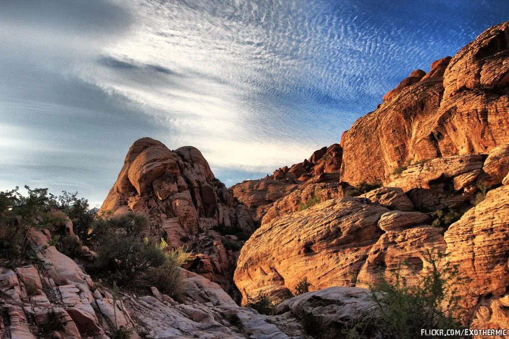 A rocky desert landscape with large reddish-brown rock formations under a blue sky with high, wispy clouds.
