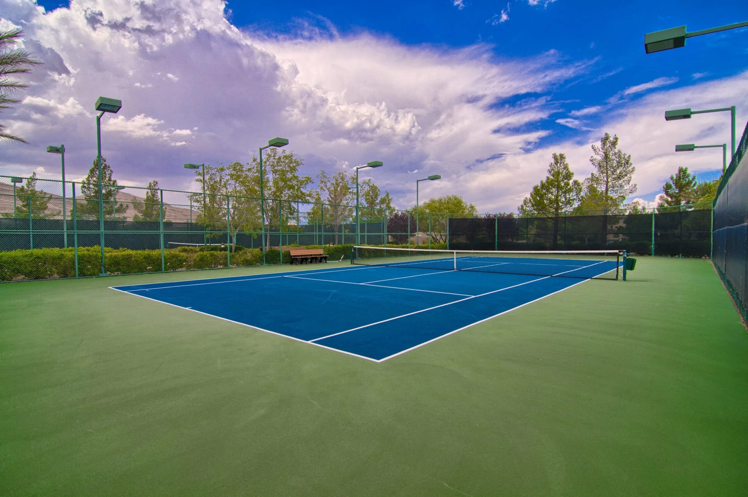 Empty Southern Highlands Golf Club outdoor tennis court with blue playing surface, surrounded by green fencing and trees under a partly cloudy sky.