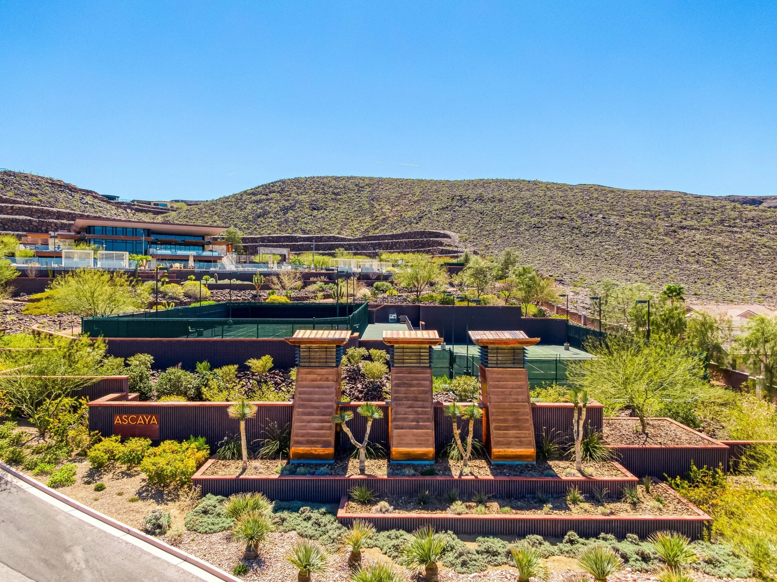 A residential community with desert landscaping, three wooden slides, and a tennis court surrounded by trees and mountains under a clear blue sky.